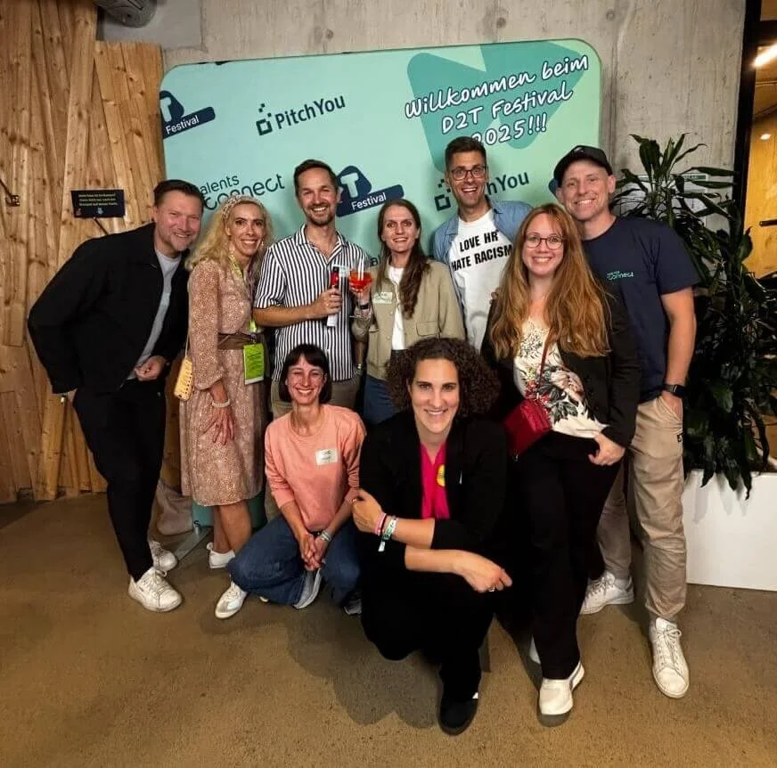 Gruppe von zehn Menschen, die bei einem Festival mit dem Motto 'Willkommen beim D2T Festival 2025' feiern, einige halten Getränke, im Hintergrund eine Begrüßungswand und Pflanzen.