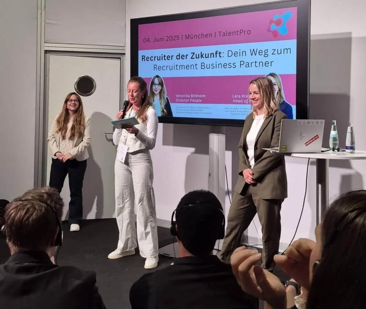 Drei Frauen bei einer Präsentation auf einer Bühne. Ein Bildschirm im Hintergrund zeigt den Titel "Recruter der Zukunft: Dein Weg zum Recruitment Business Partner" auf Deutsch. Das Publikum sitzt vor der Bühne.