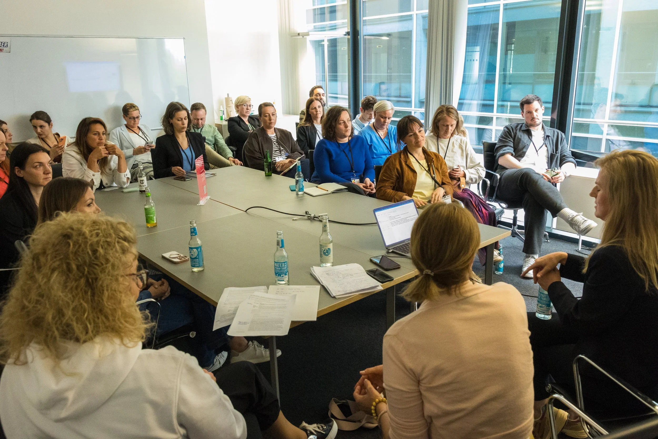 Menschen sitzen in einem modernen Büro-Meetingraum mit großen Fenstern, einige schauen auf ihre Smartphones, andere hören aufmerksam den Rednern zu.