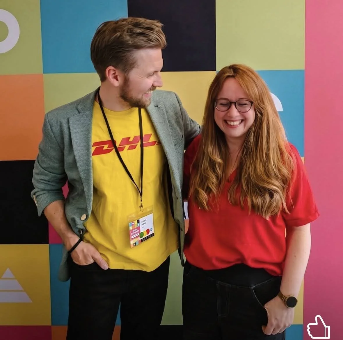 Ein Mann und eine Frau lachen und stehen nebeneinander vor einem bunte Wand. Der Mann trägt ein graues Sakko und ein gelbes DHL T-Shirt, die Frau trägt eine rote Bluse und eine Uhr am Handgelenk.