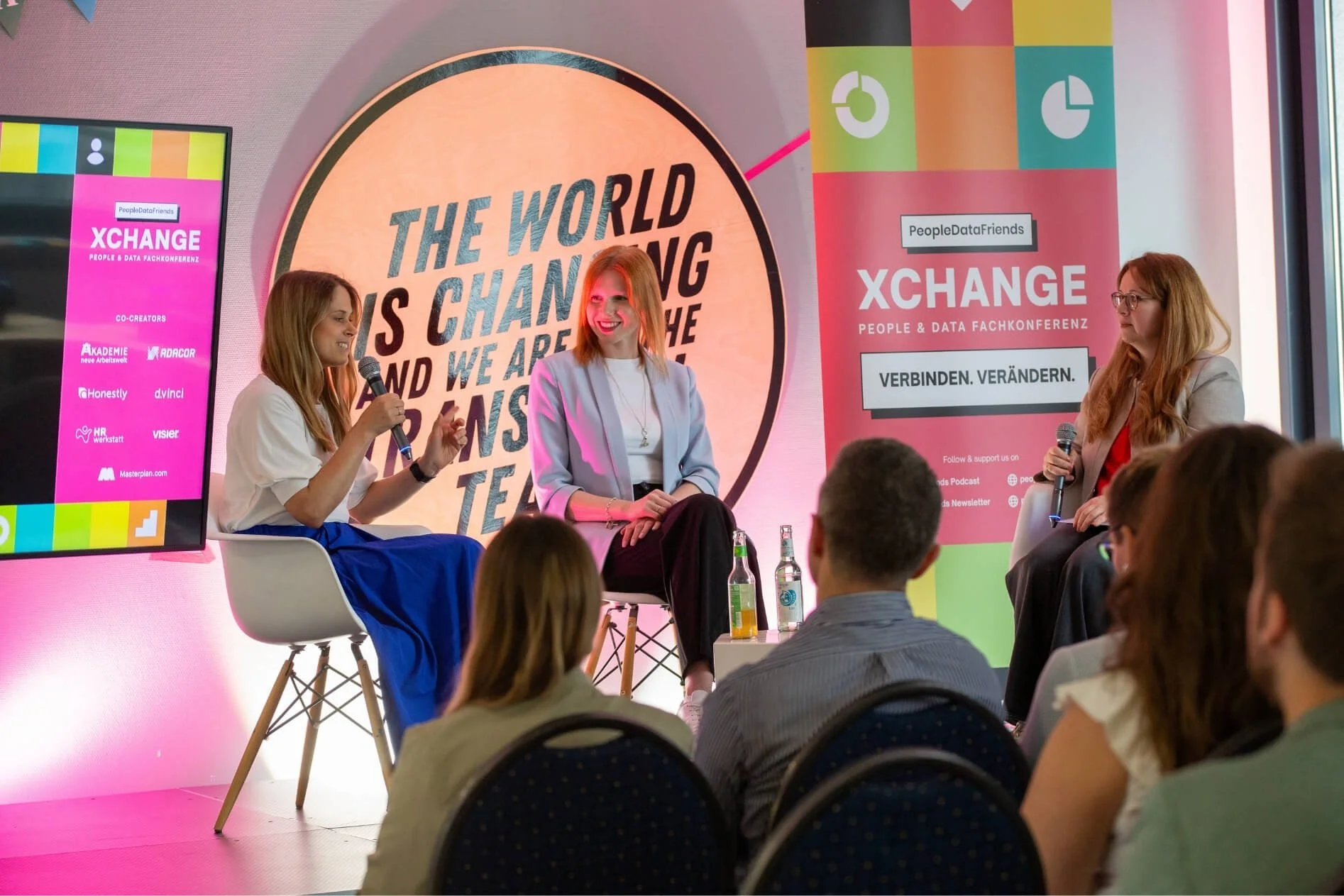 Drei Frauen diskutieren bei einer Konferenz namens XCHANGE. Es gibt ein großes Display im Hintergrund mit der Botschaft 'The world is changing and we are the change'. Die Damen sitzen auf Stühlen und halten Mikrofone, während das Publikum zuhört.