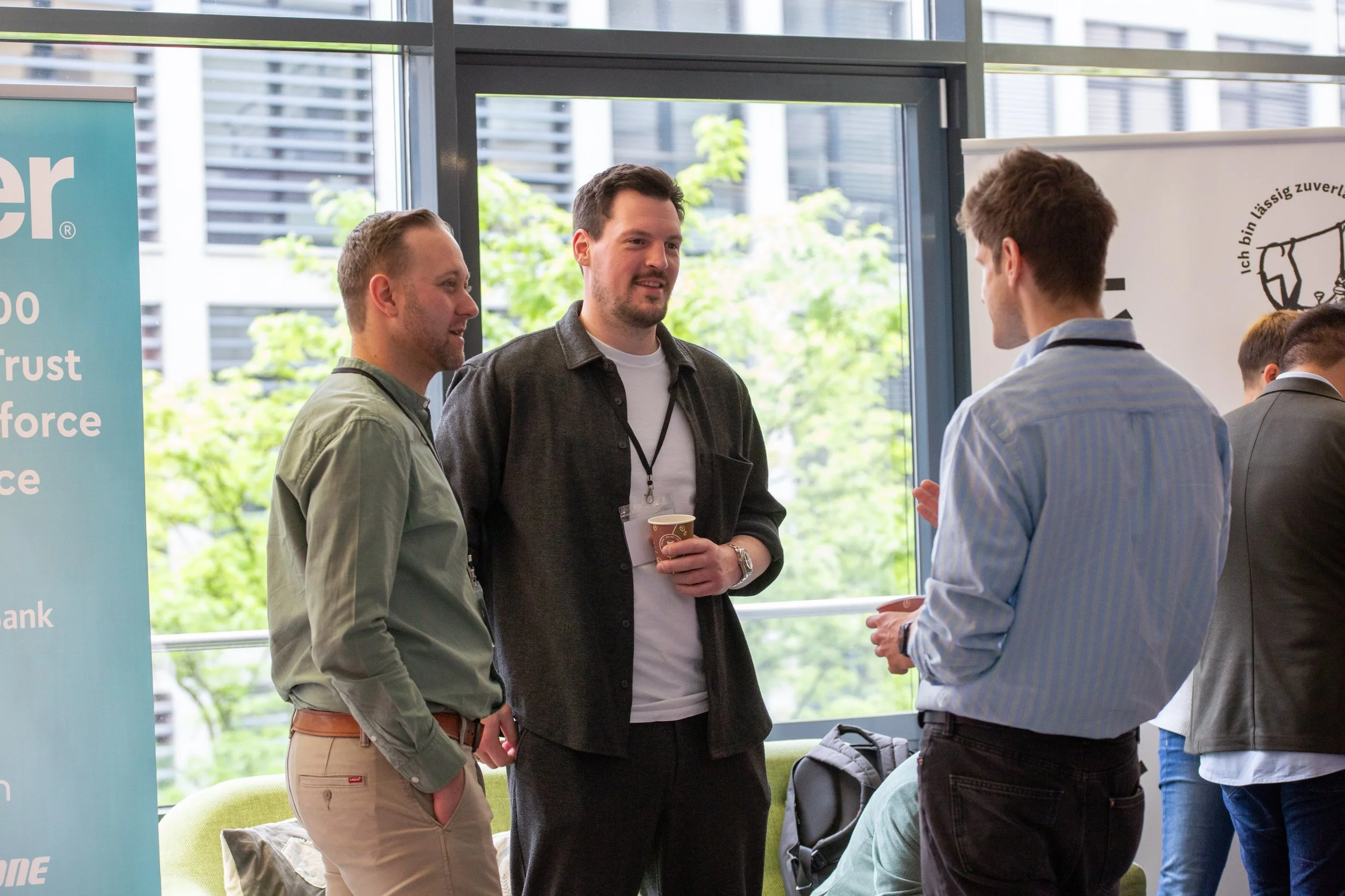 Drei Männer im Gespräch bei einer Konferenz oder einem Networking-Event im Raum mit großen Fenstern. Einer hält eine Kaffeetasse, während sie sich unterhalten.