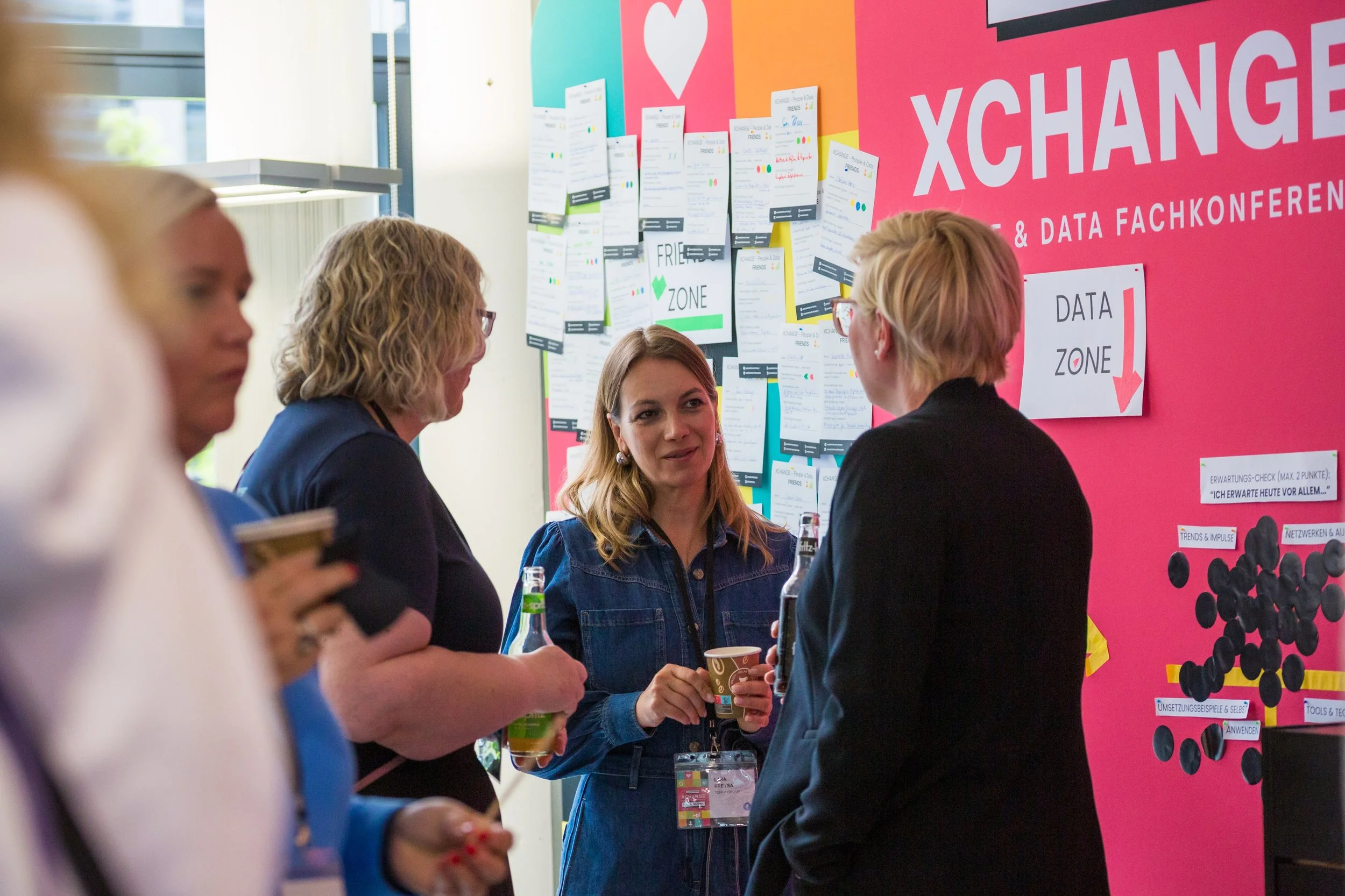 Menschen bei einer Fachkonferenz, im Hintergrund ein pinker Präsentationswand mit der Aufschrift 'XCHANGE' und mehreren beschrifteten Pinnwänden mit Notizen.