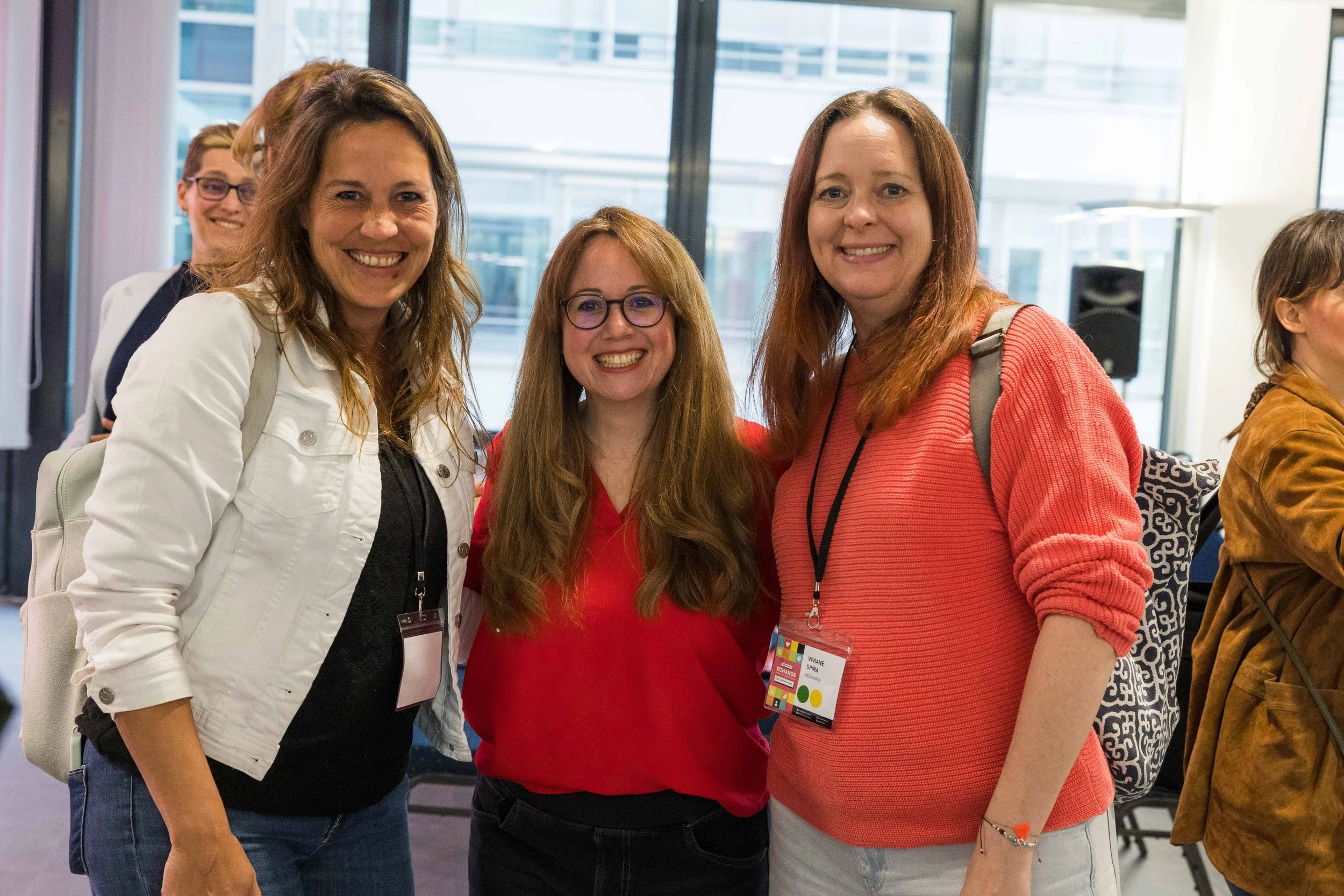 Vier Frauen lächeln in einem modernen, hellen Innenraum mit großen Fenstern, beim Konferenz- oder Networking-Event. Zwei der Frauen tragen Namensschilder, eines ist schwarz mit bunten Akzenten.