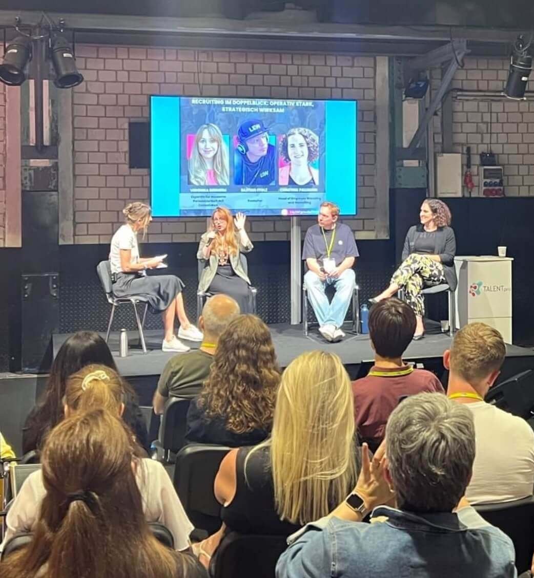Bühne mit vier Personen, ein Bildschirm im Hintergrund zeigt ein Panel mit dem Titel „Security in DoppelRück: Operative Stamm, Strategisch Wirksam“, und im Publikum sitzen diverse Menschen.