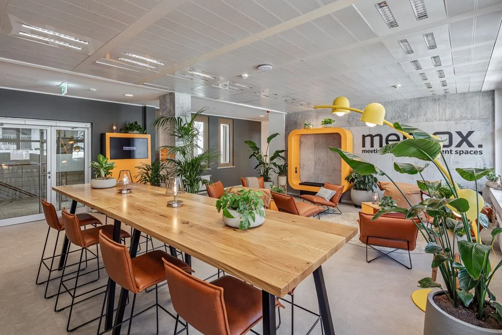 Moderner Gemeinschaftsbereich mit Holztisch, braunen Stühlen, grünen Pflanzen und einer Lounge-Ecke mit orange Möbel und gelbem Stehlampenschirm.