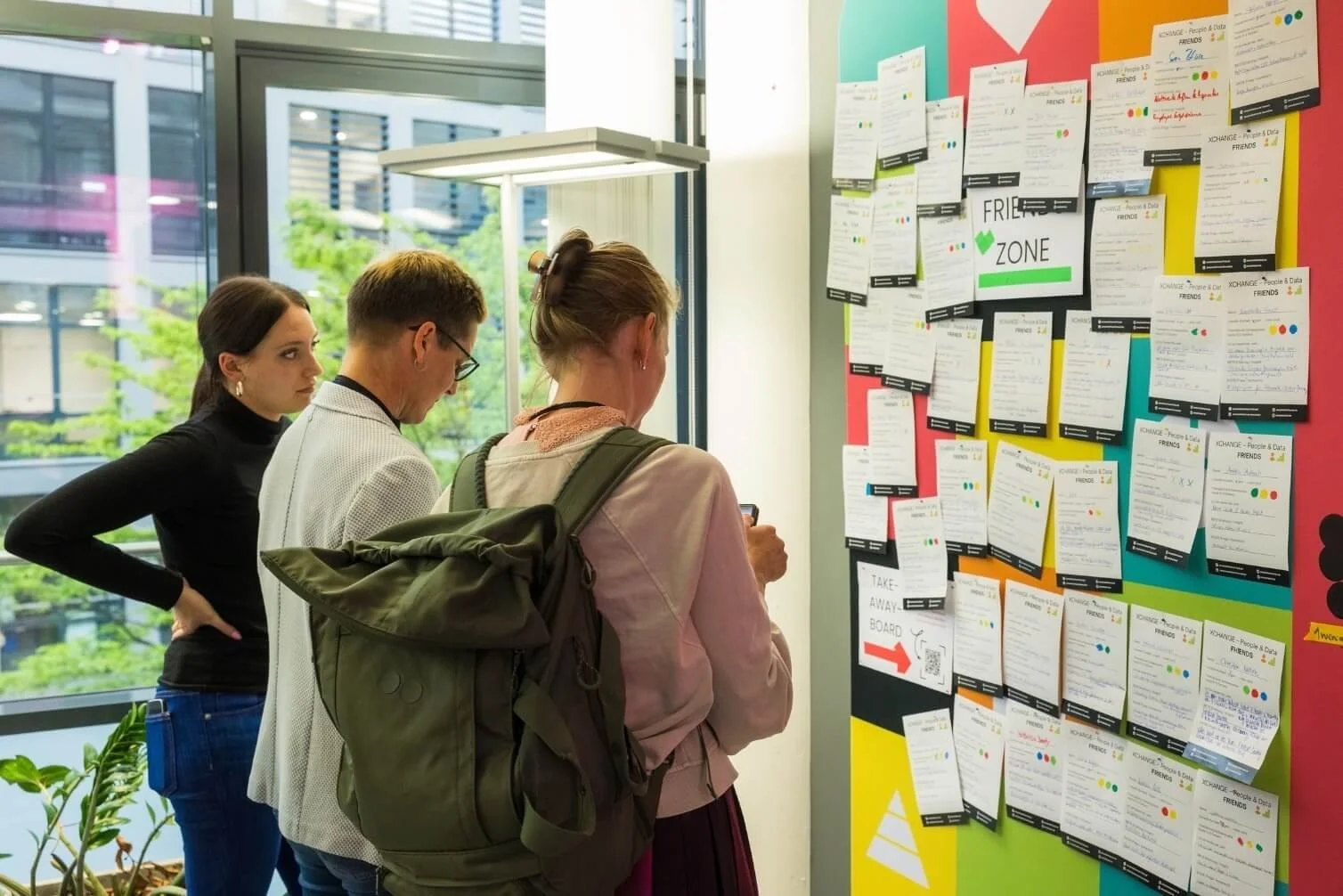 Drei Menschen lesen aushänge an einer Pinnwand in einem modernen Gebäude, wahrscheinlich eine Schule oder ein Gemeinschaftszentrum.