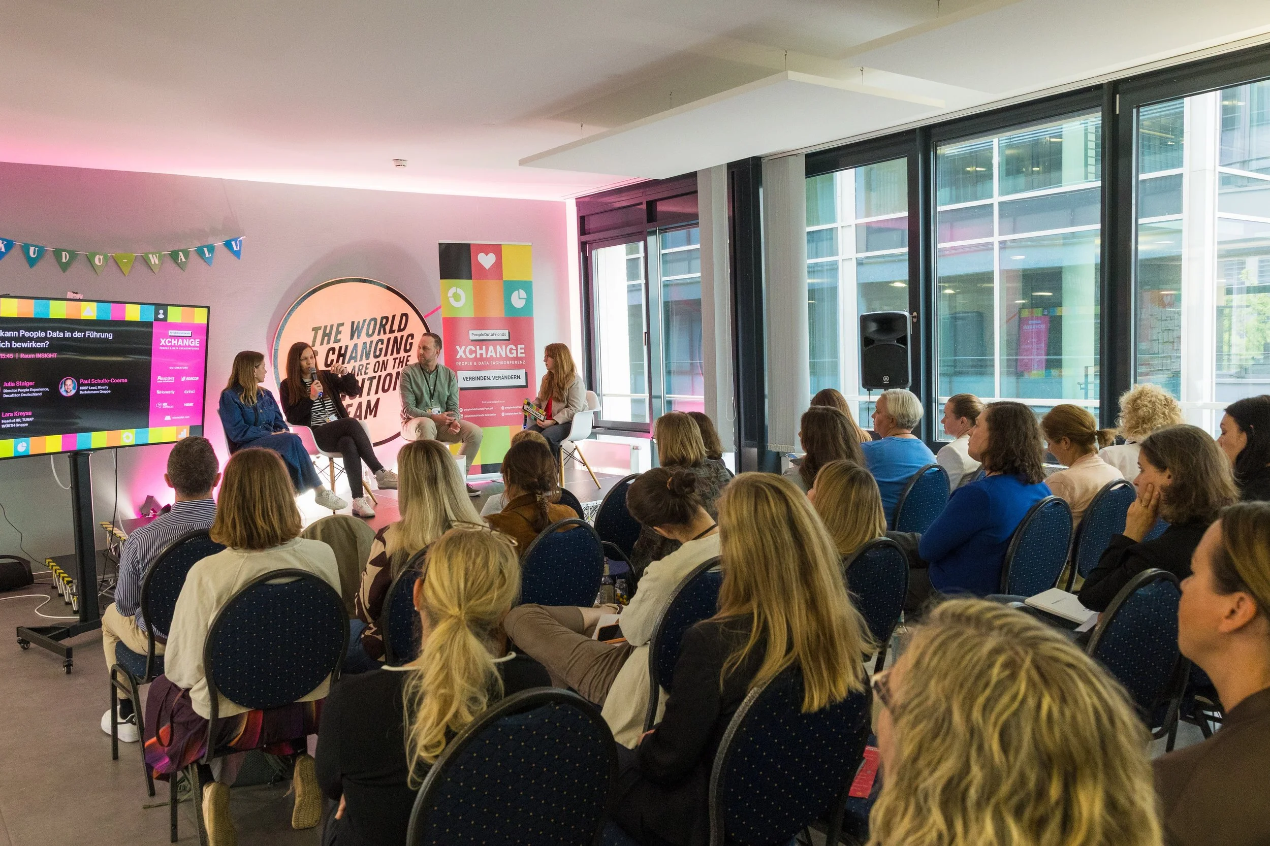 Ein Panel bei einer Konferenz mit vier Personen, die auf der Bühne sitzen, und einem Publikum, das zuhört. Es gibt eine große Anzeige mit bunten Farben und Schrift, und eine Wanddekoration mit bunten Dreiecken.