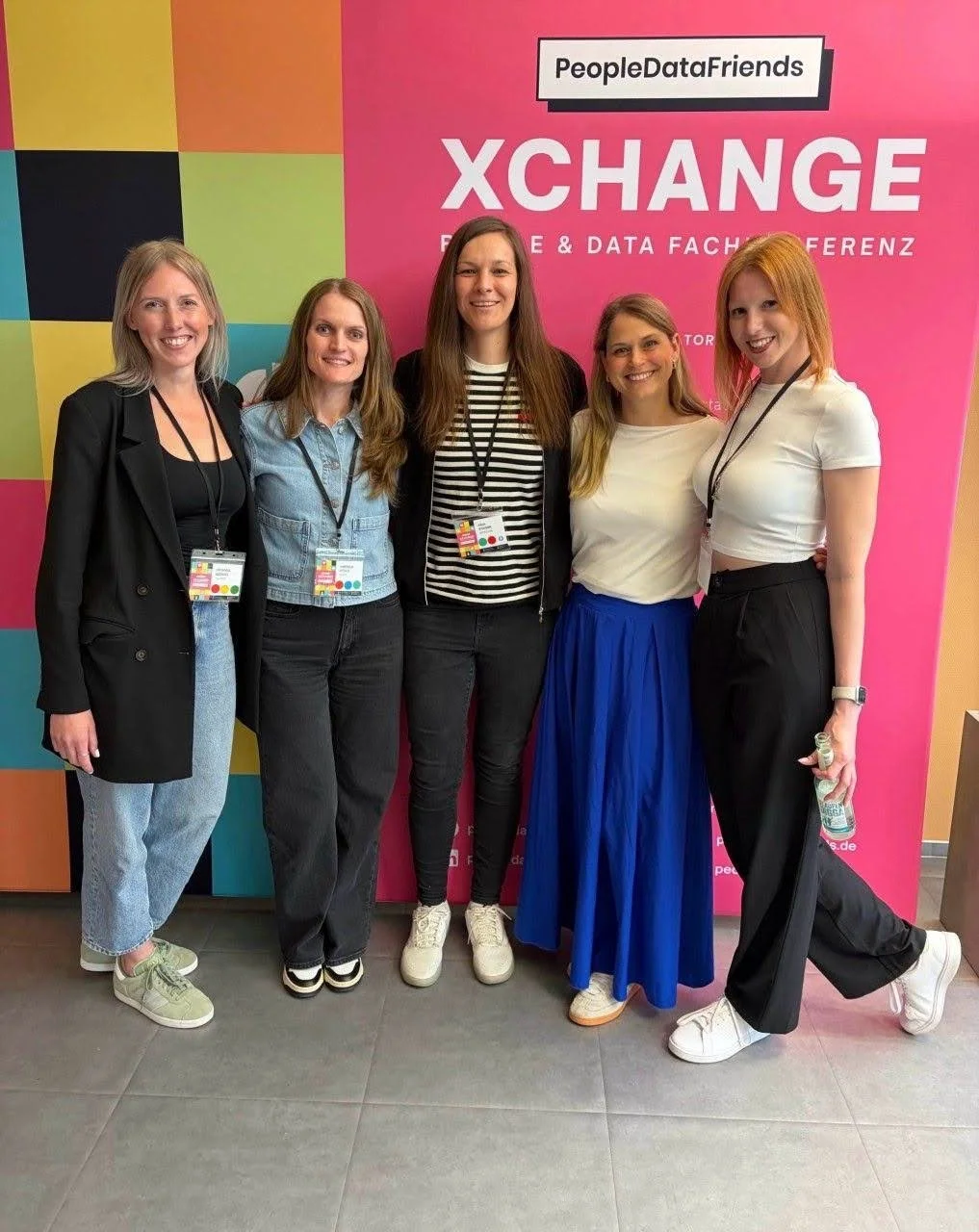 Fünf Frauen stehen lächelnd nebeneinander vor einem bunten Hintergrund mit der Aufschrift 'PeopleDataFriends XCHANGE'. Sie tragen Konferenzbänder um den Hals, und eine Frau auf der rechten Seite hält eine Wasserflasche.