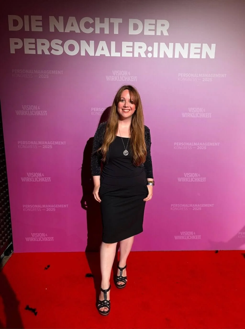 Frau in schwarzem Kleid mit lange Haaren, steht auf rotem Teppich vor pinkem Hintergrund mit Texten, lächelt.