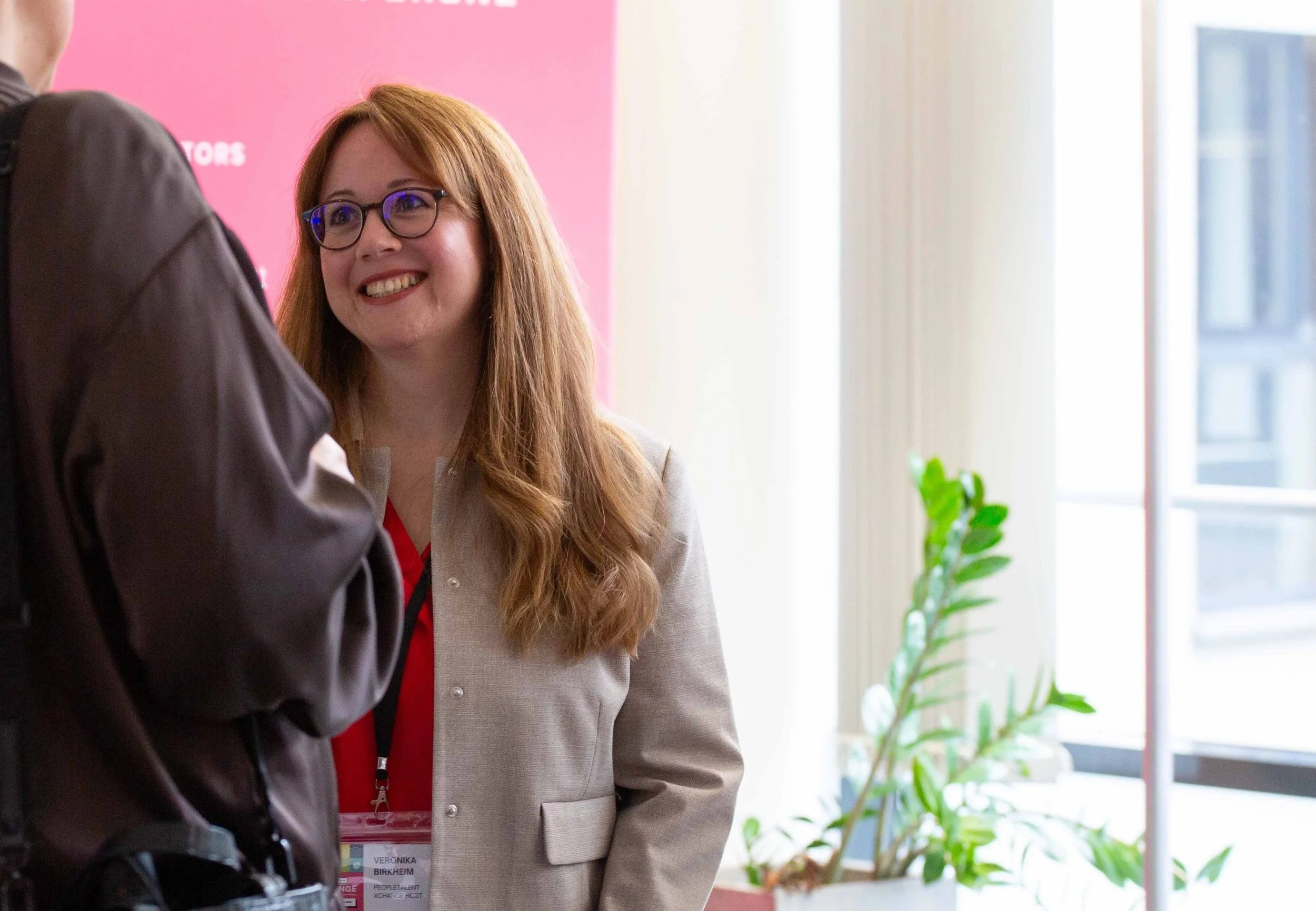 Eine lachende Frau mit roten Haaren und Brille führt ein Gespräch mit einem Mann. Sie trägt eine beige Jacke und hat einen Namensschild mit ihrem Namen und ihrer Funktion. Im Hintergrund gibt es ein Fenster und eine grüne Pflanze.