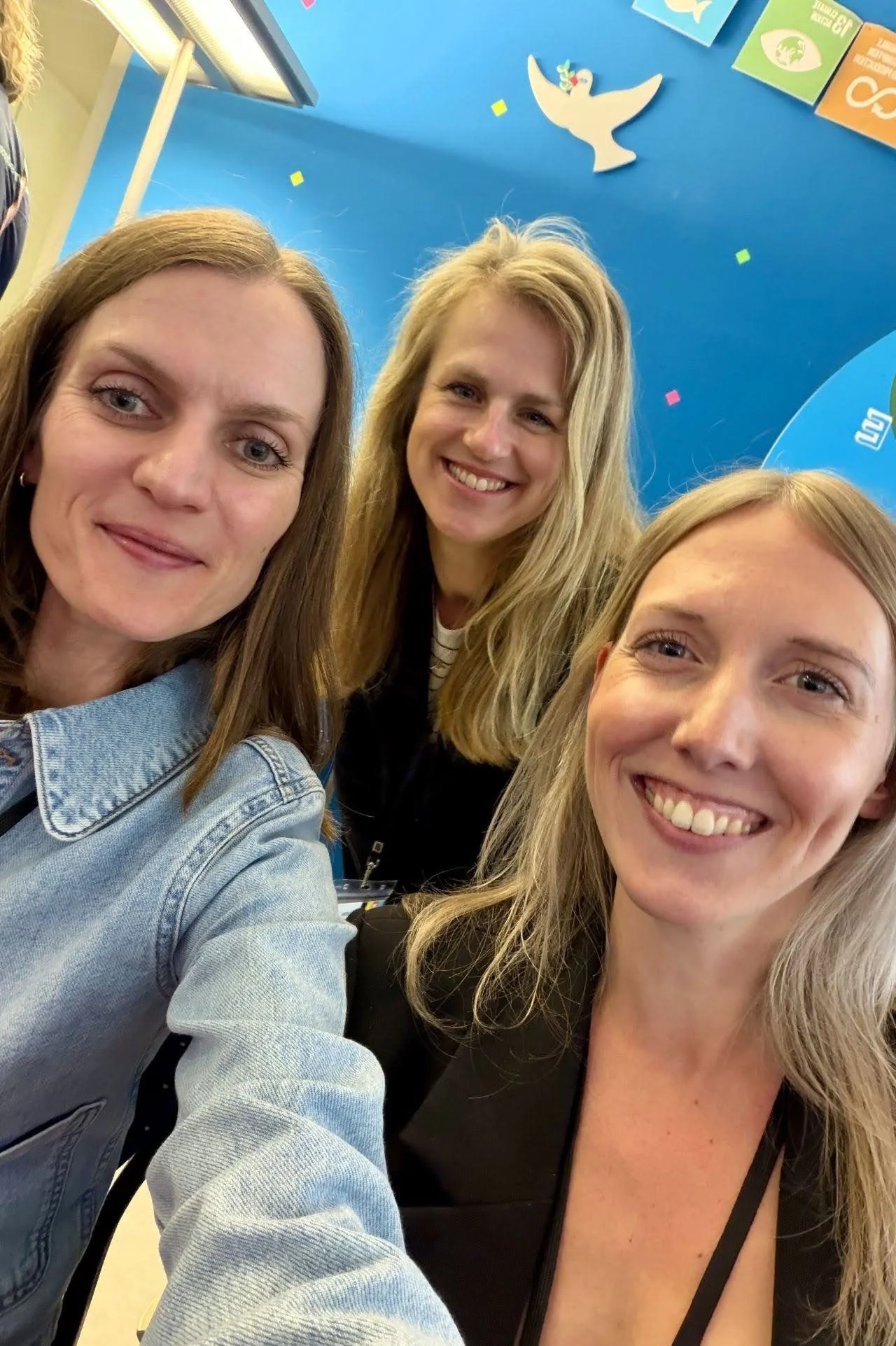 Drei Frauen nehmen ein Selfie vor einer blauen Wand mit einer weißen Taube und bunten Symbolen. Alle lachen freundlich.