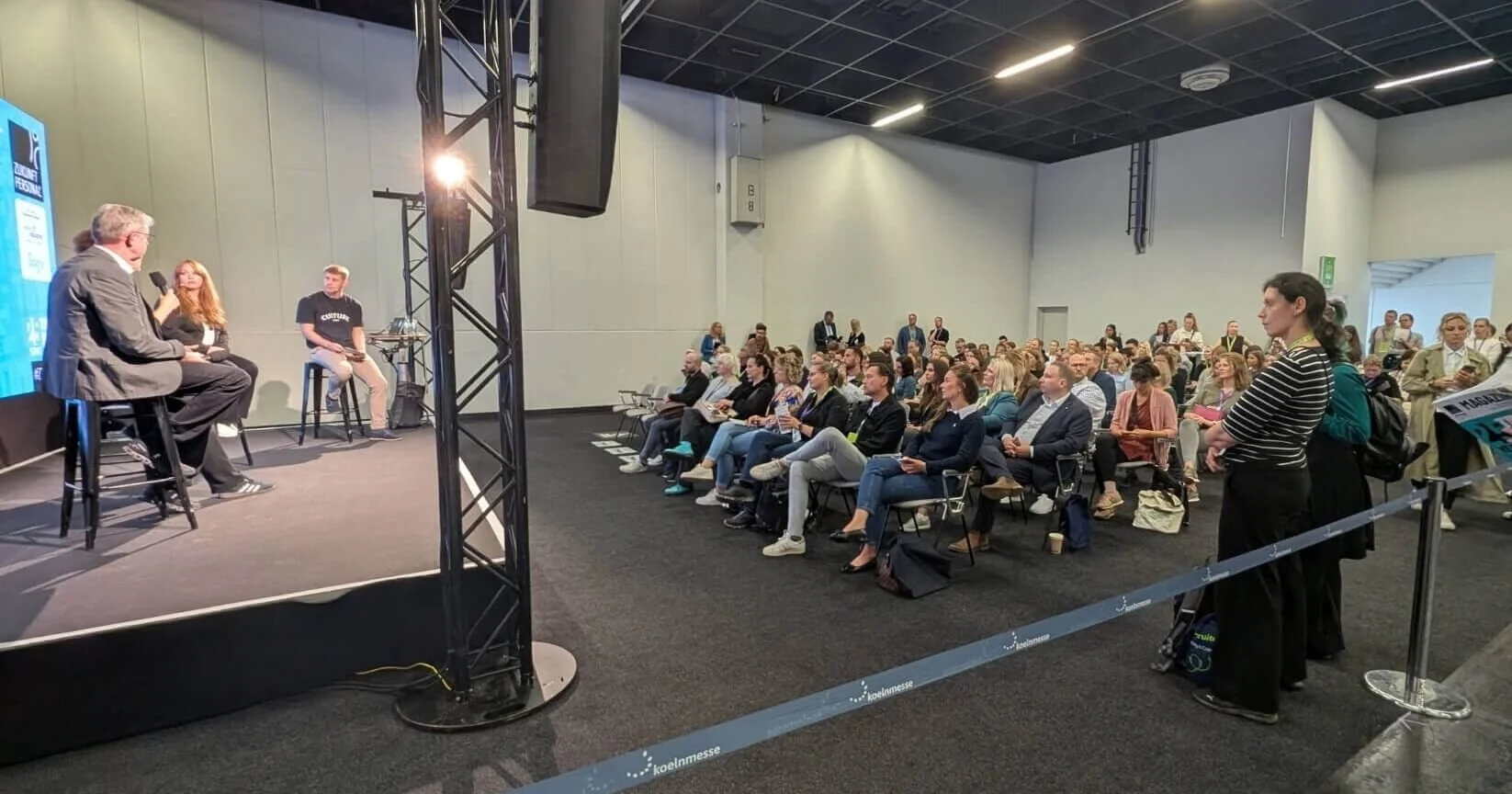 Eine Gruppe von Menschen sitzt in einem verschlossenen Saal und hört einem Gespräch auf der Bühne zu, auf der drei Personen sitzen, die an einer Diskussion teilnehmen.