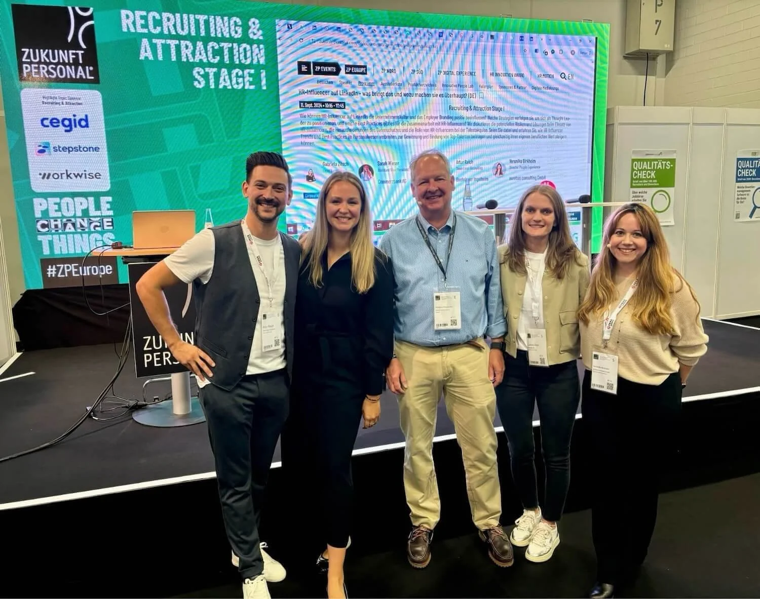 Fünf Menschen stehen nebeneinander vor einem großen, grünen Bildschirm auf einer Messe, alle lächeln. Der Bildschirm zeigt Text und Logos im Bereich für Recruiting und Personal. Die Gruppenmitglieder tragen Namensschilder und sind in informeller Busi