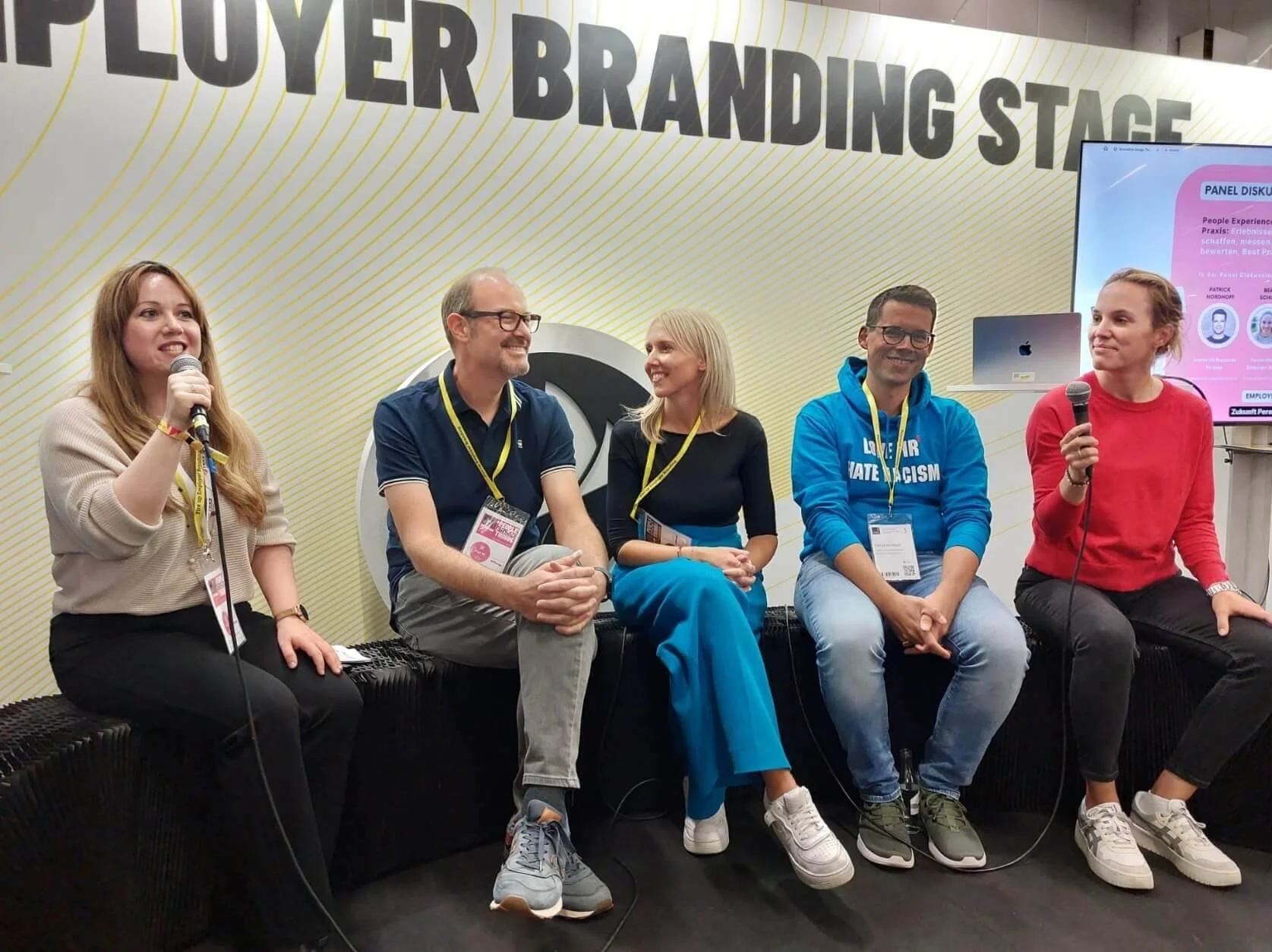 Fünf Menschen sitzen auf einer Bank auf einer Bühne bei einer Podiumsdiskussion. Zwei Frauen auf der linken und rechte Seite halten Mikrofone. Im Hintergrund steht eine große Tafel mit der Aufschrift 'LOYER BRANDING STAGE'.