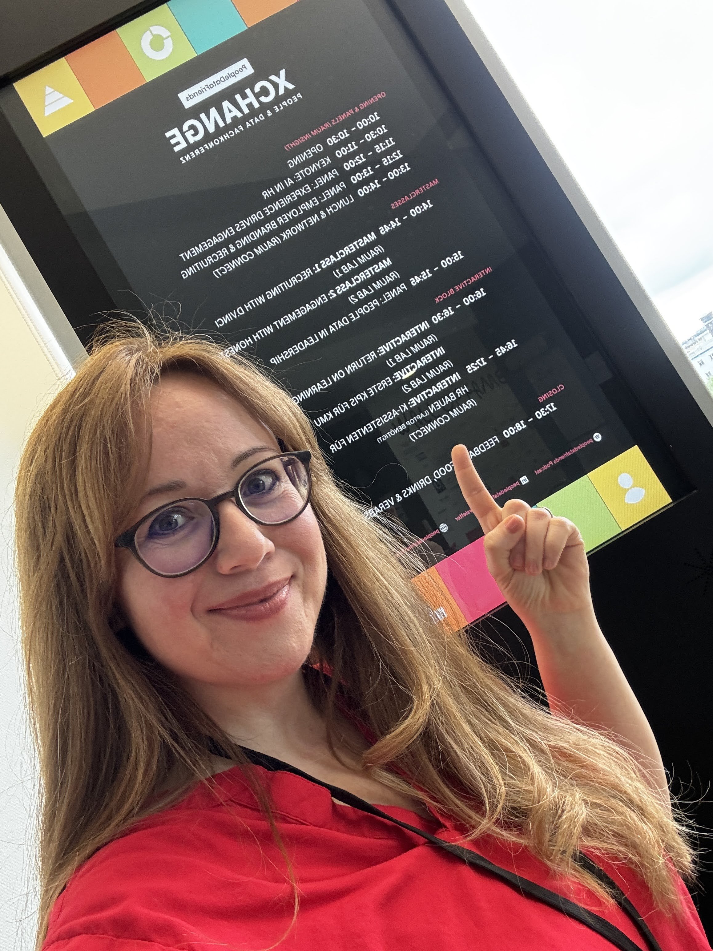 Frau mit Brille hält Finger in die Luft, vor einem großen Bildschirm mit Programmübersicht, im Büro.