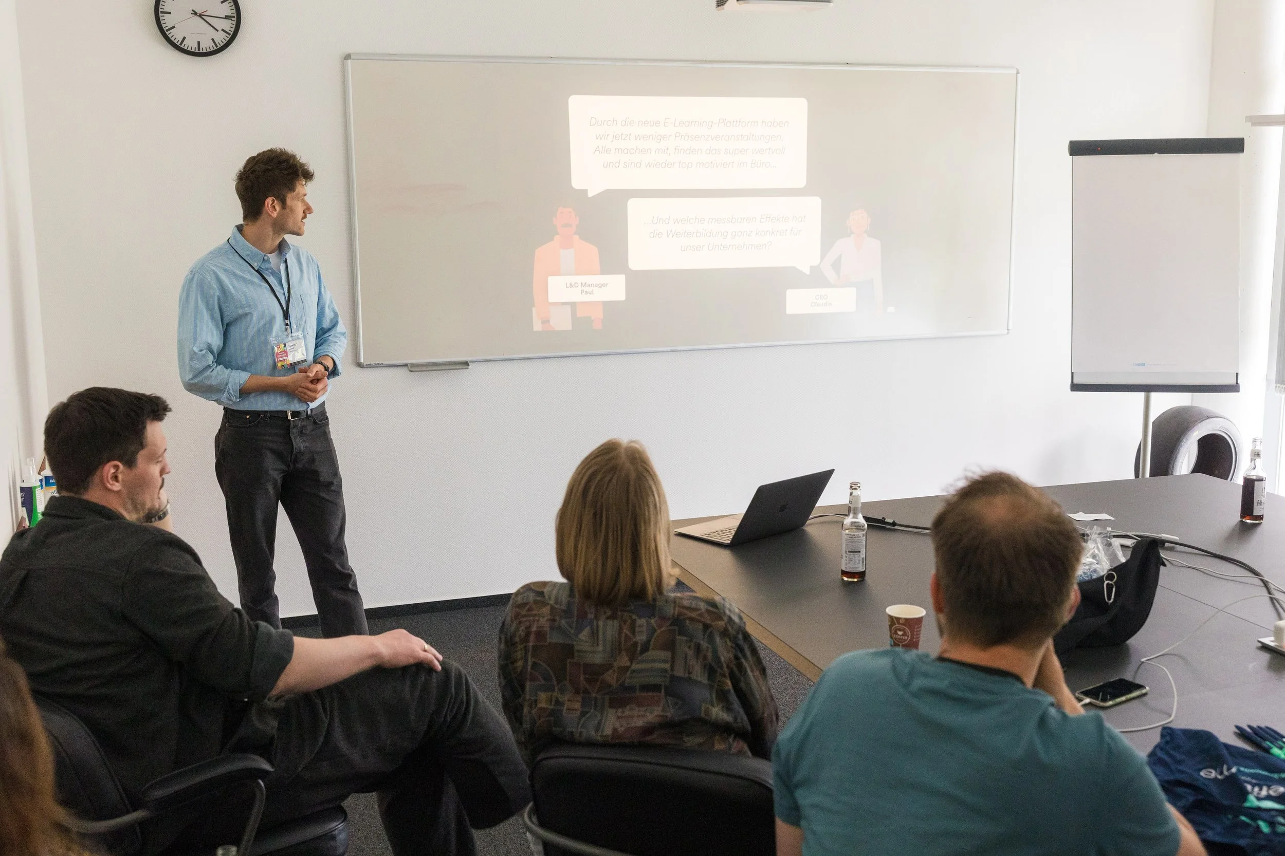 Eine Gruppe von Menschen sitzt in einem Konferenzraum und hört einem Mann zu, der vorne steht. Es gibt einen weißen Whiteboard, eine Projektionswand mit Text, einen Laptop auf dem Tisch, Wasserflaschen und Kaffeetassen.