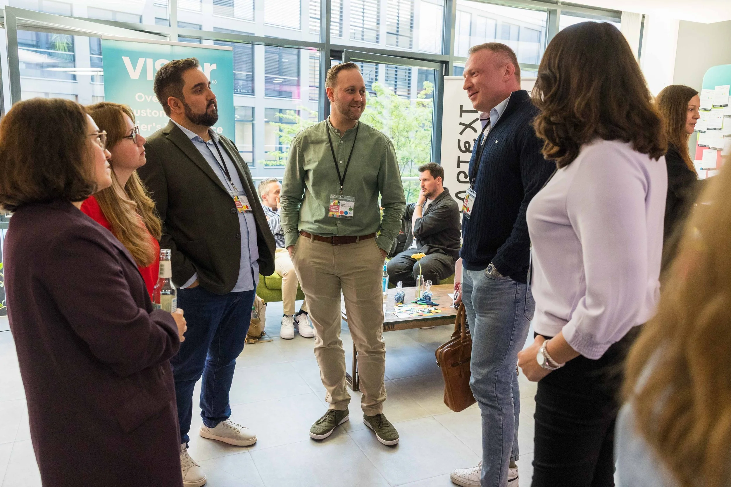 Gruppengespräch bei einer Konferenz in einem hellen Raum mit großem Fenster. Mehrere Personen stehen im Kreis und unterhalten sich, im Hintergrund sind Stühle und Tische zu sehen.