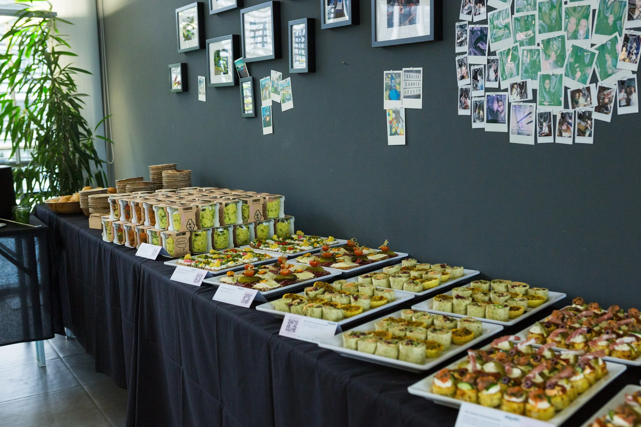 Buffet mit verschiedenen Mini-Sandwiches und Häppchen, im Hintergrund eine Wand mit Fotos und Postern, links eine Pflanze, und Tische mit Geschirr und Servierten.