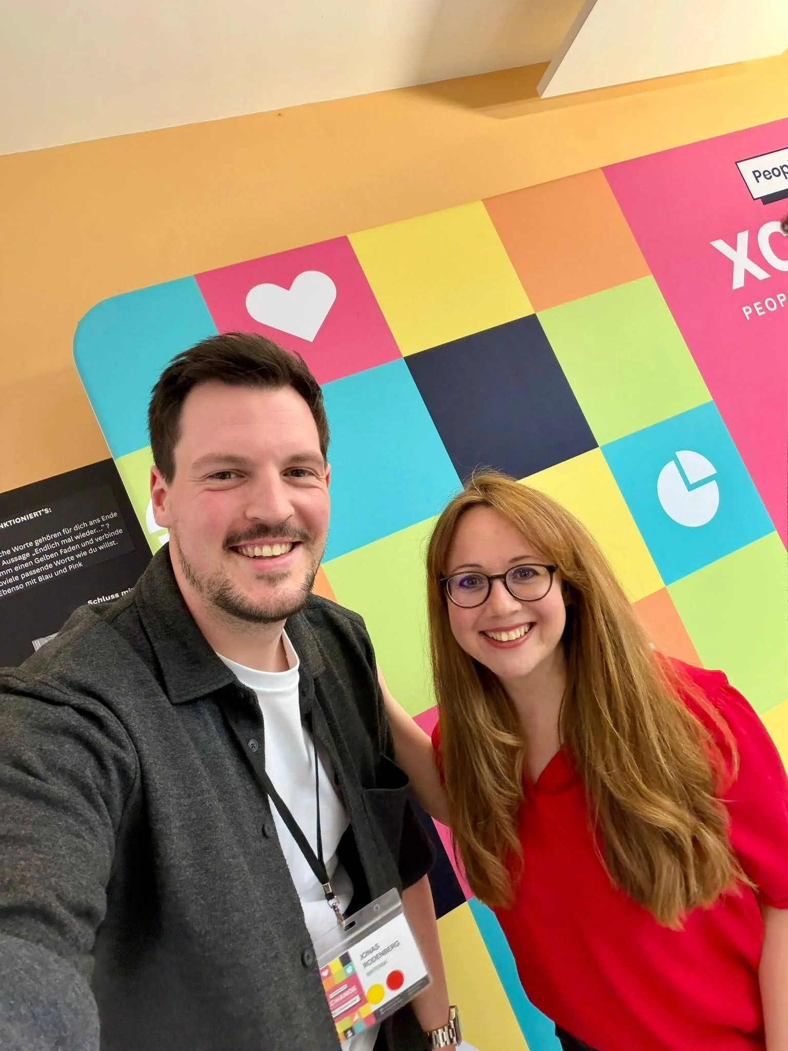 Zwei Personen, ein Mann mit dunklen Haaren und Bart, und eine Frau mit roten Haaren und Brille, posieren vor einem bunten Hintergrund mit Farbblöcken und Symbolen bei einer Konferenz oder Veranstaltung.