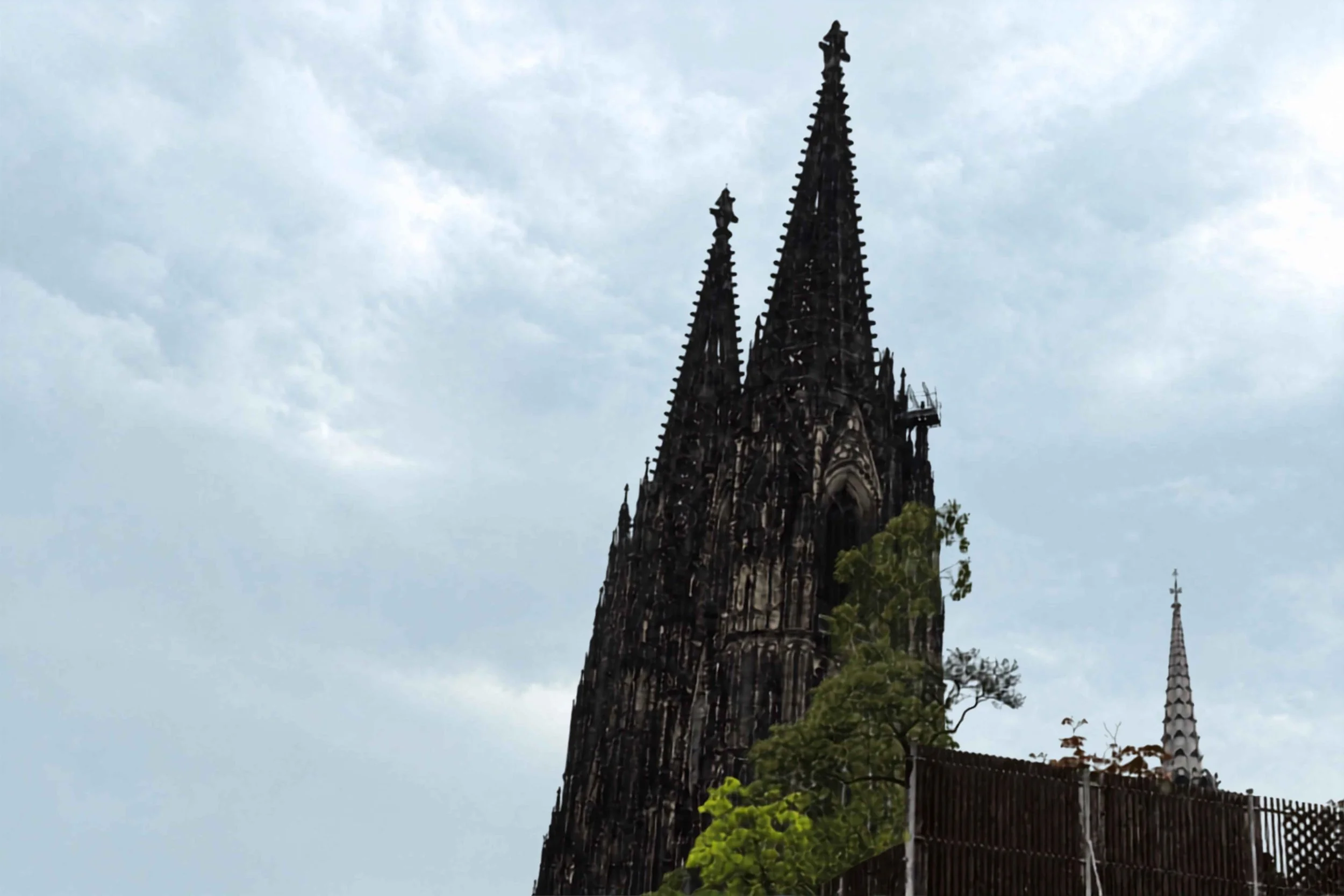 Der romanische Kirchturm mit Spitzdach und Zinnen im Himmel, umgeben von Bäumen und einer Zäunung.