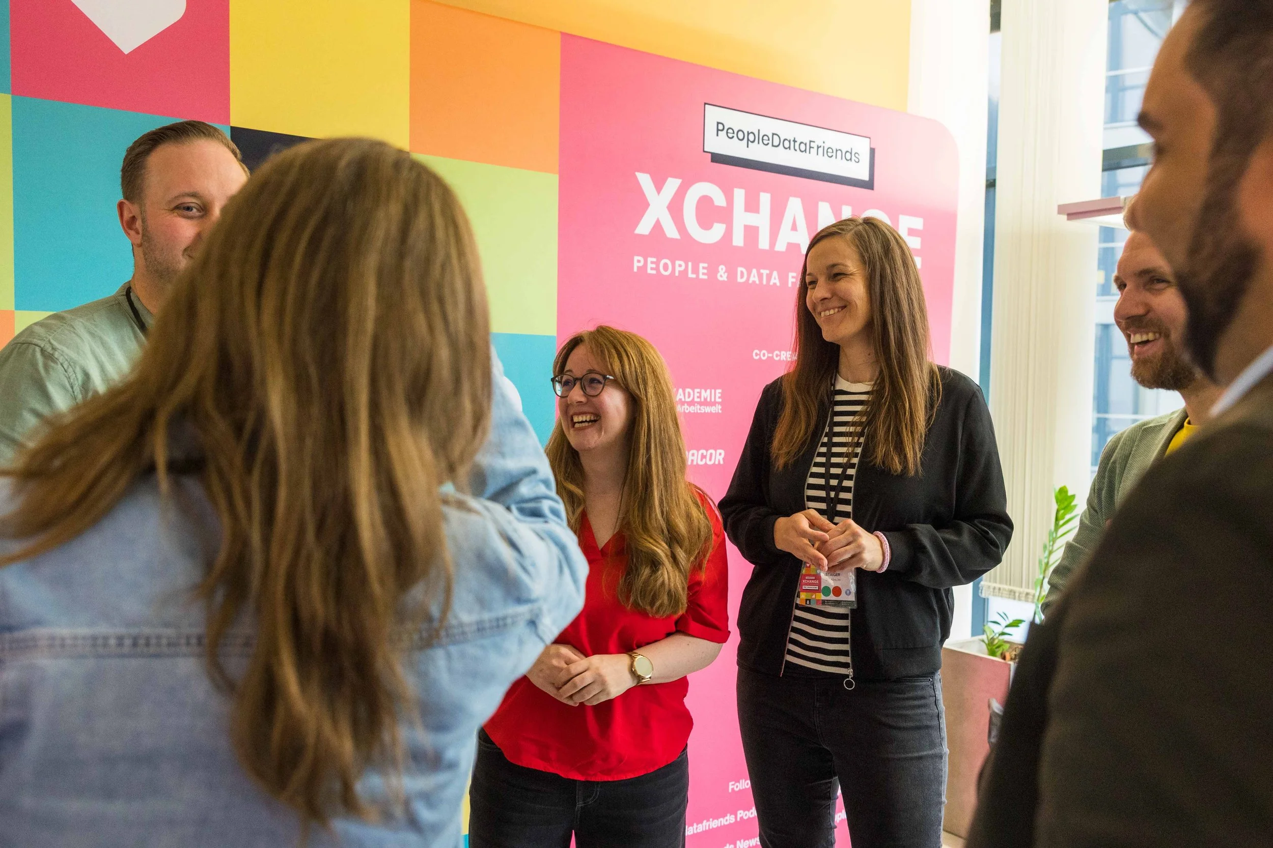Mehrere Menschen beim Gespräch bei einer Austauschveranstaltung mit einem pinken Banner im Hintergrund, das 'XCHANGE' und 'People & Data Friends' schreibt.
