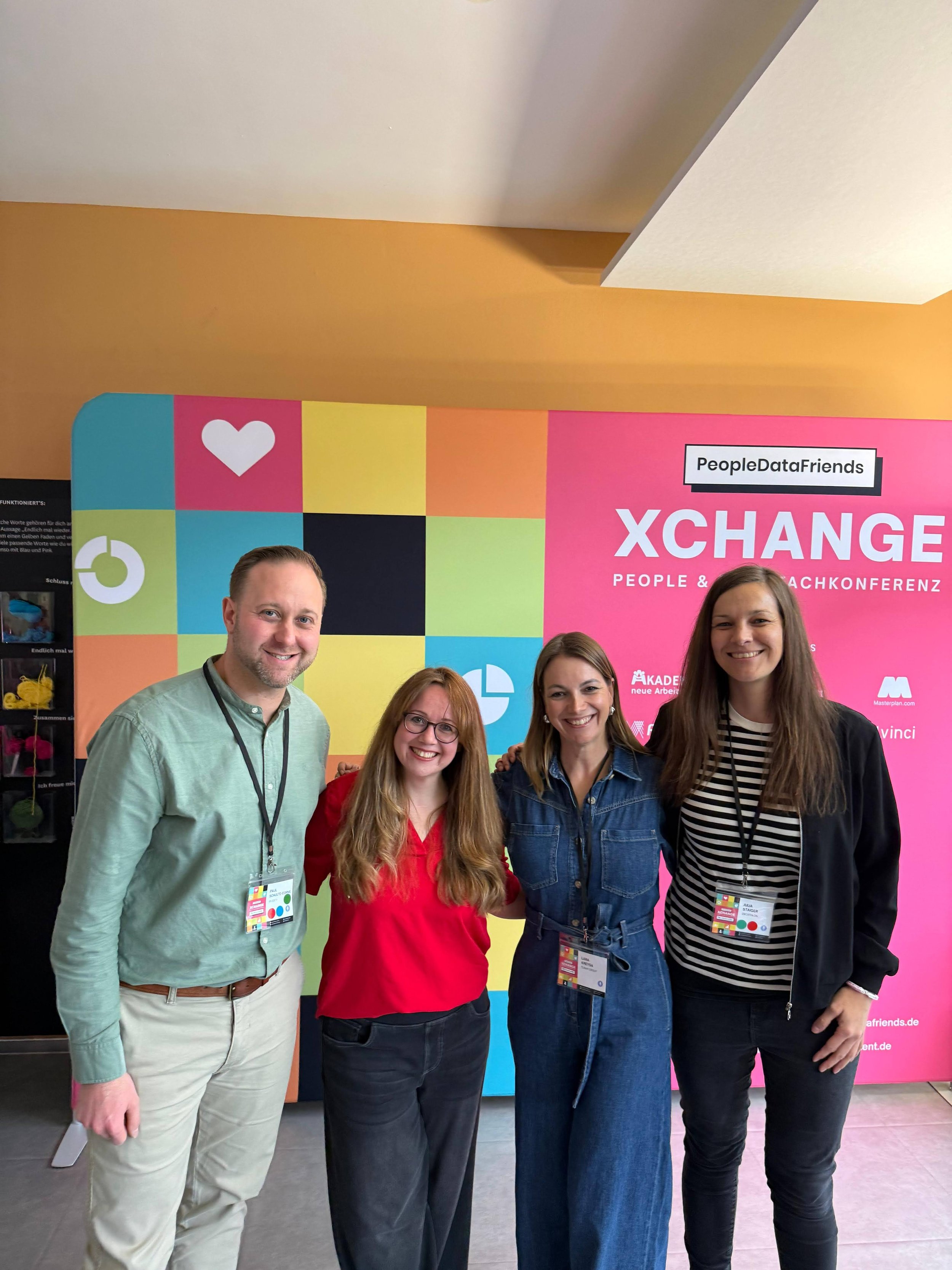 Fünf Menschen stehen vor einem bunten Messe-Hintergrund mit der Aufschrift 'XCHANGE' und 'People & Strategie Konferenz'.