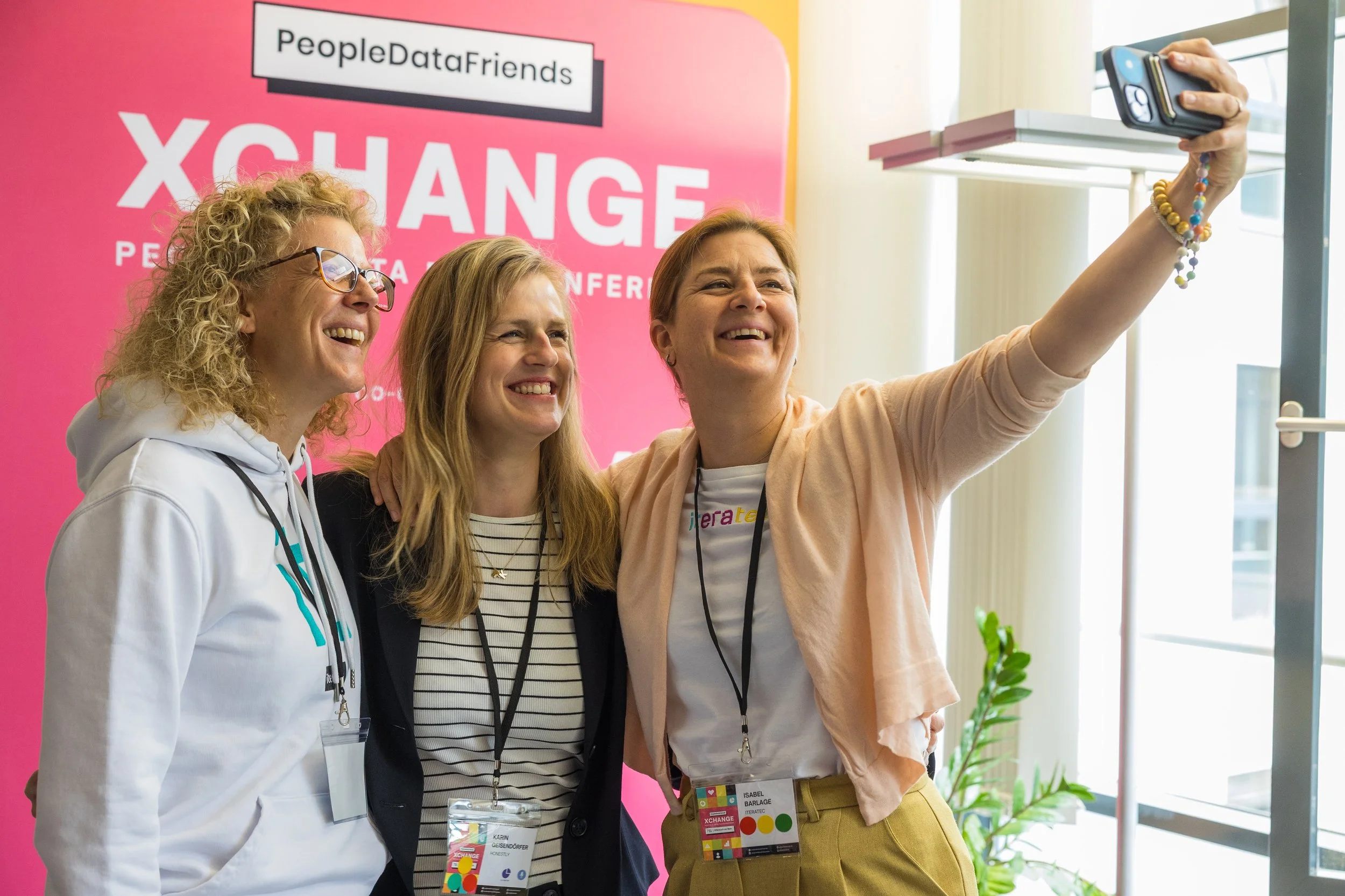 Drei Frauen nehmen ein Selfie vor einem pinken Hintergrund mit dem Text 'PeopleDataFriends XCHANGE' auf einer Konferenz. Sie lachen und posieren freudig.