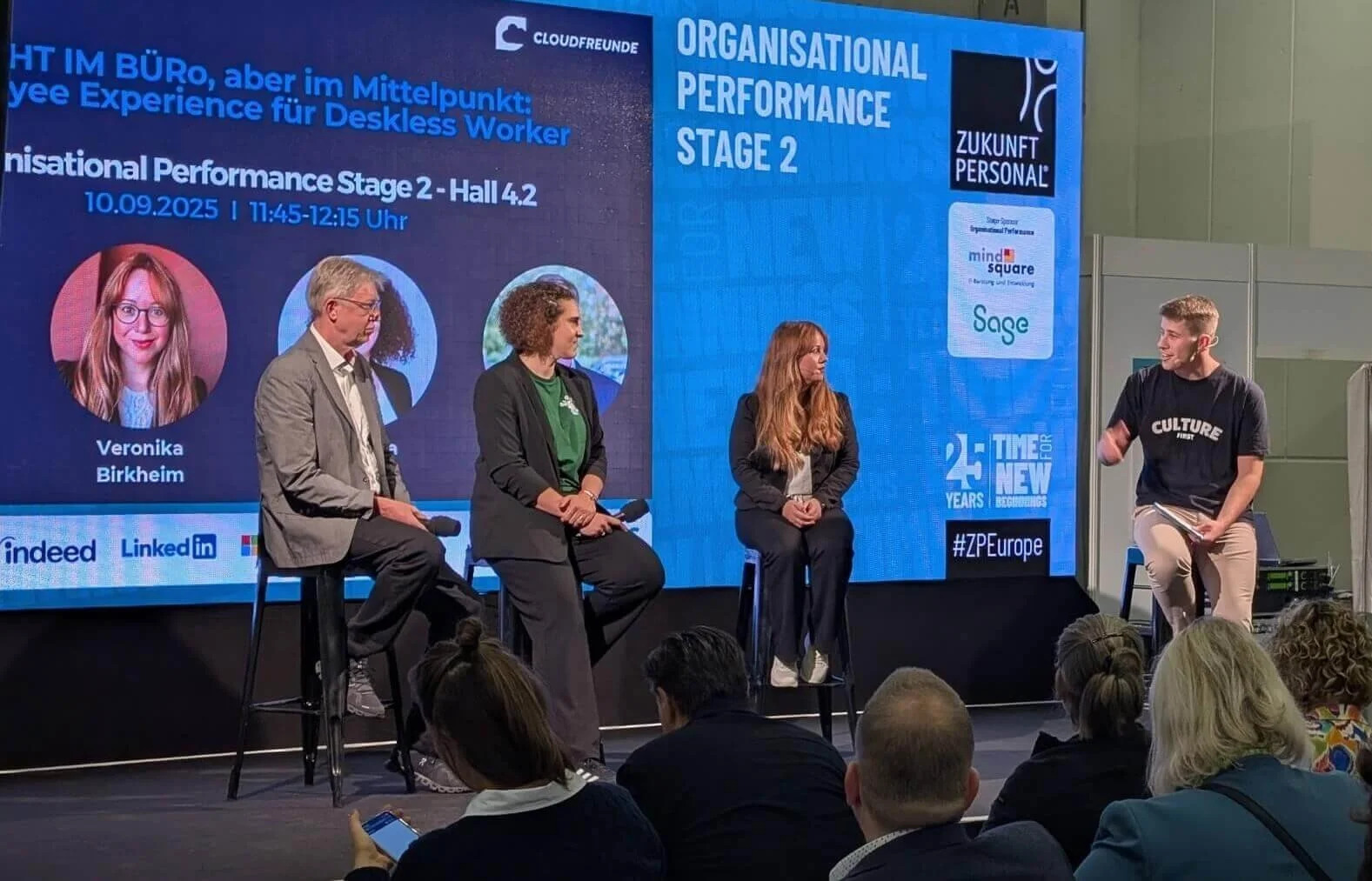 Bühne mit einer Panel-Diskussion während einer Konferenz, drei Frauen und ein Mann sitzen auf Stühlen, eine Person steht rechts und spricht. Der Hintergrund zeigt einen großen Bildschirm mit Veranstaltungsinformationen, Logos und Text auf Deutsch und