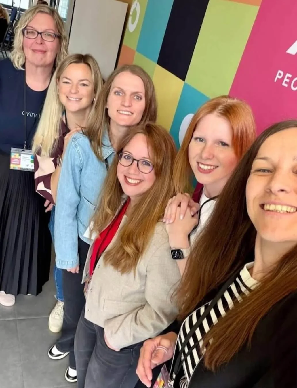 Gruppenfoto von sechs Frauen, die bei einer Veranstaltung mit buntem Hintergrund stehen und lächeln.