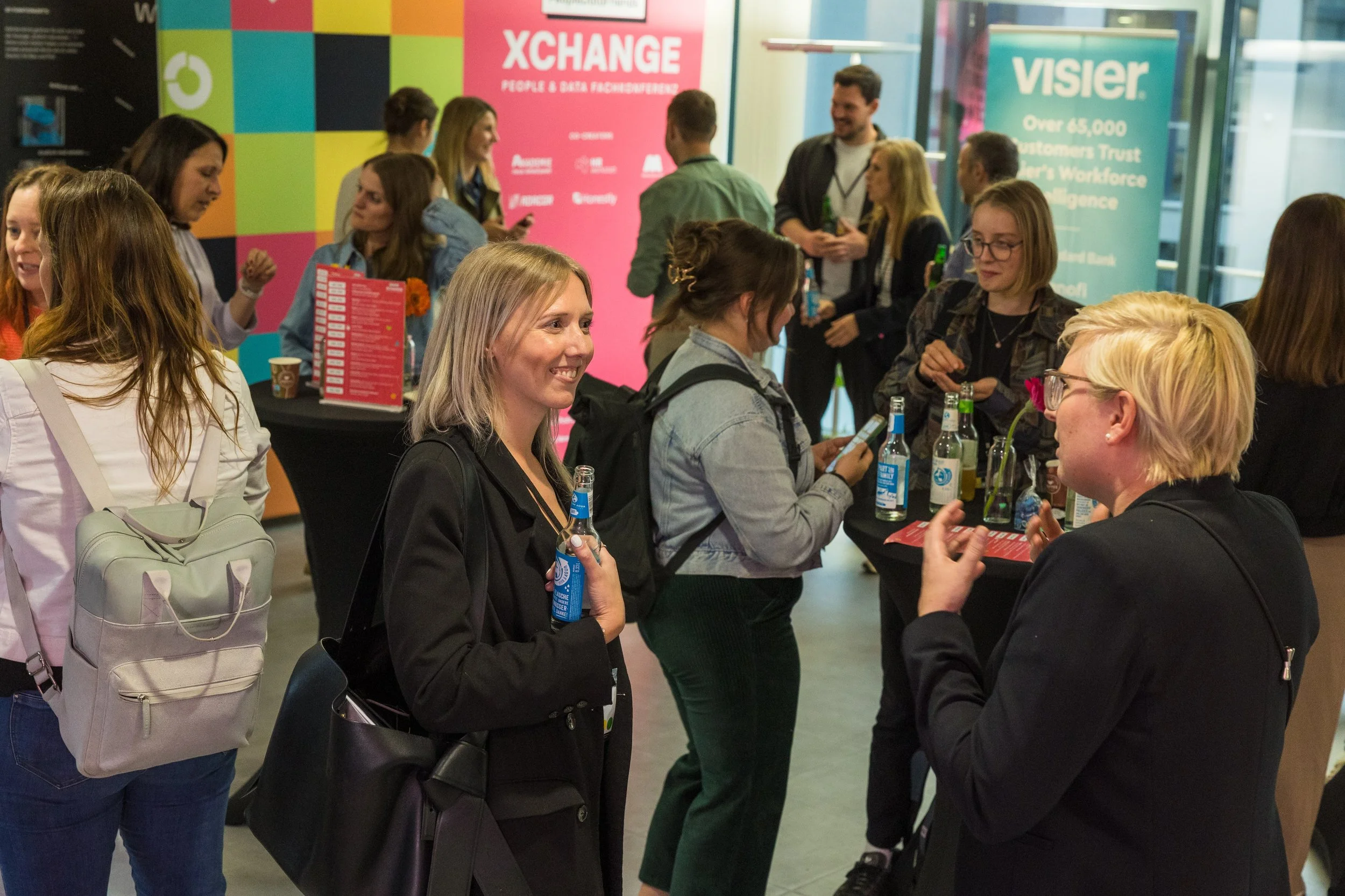Menschen bei einer Networking-Veranstaltung, einige sitzen an einem Tisch und sprechen, andere stehen im Hintergrund, bunte Veranstaltungsbanner sind sichtbar.