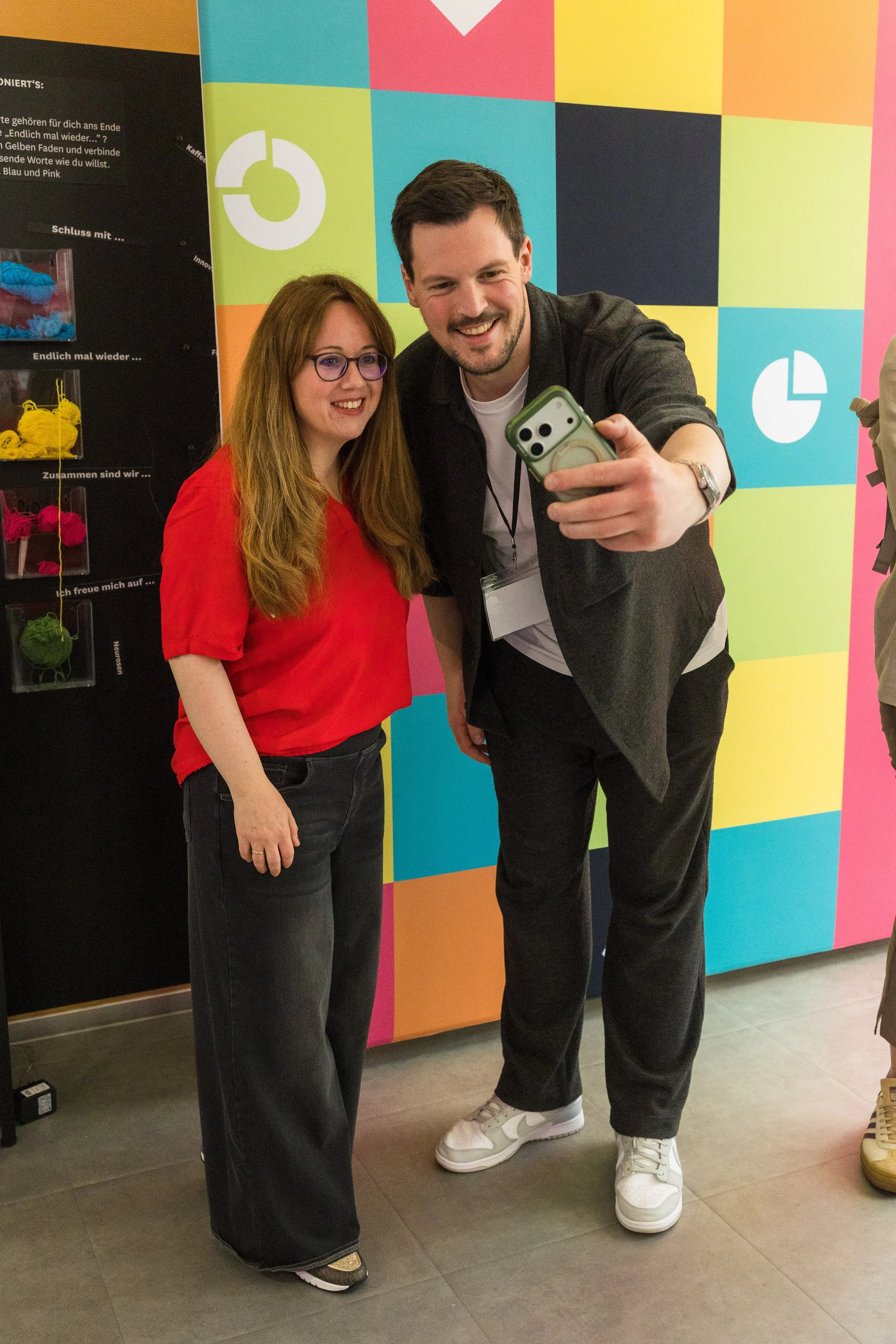Zwei Menschen, ein Mann und eine Frau, nehmen ein Selfie vor einem bunten, quadratischen Wandbild auf. Der Mann trägt ein schwarzes Jackett, weiße Schuhe, und hält ein Smartphone in der Hand. Die Frau trägt eine rote Bluse und dunkle Jeans.