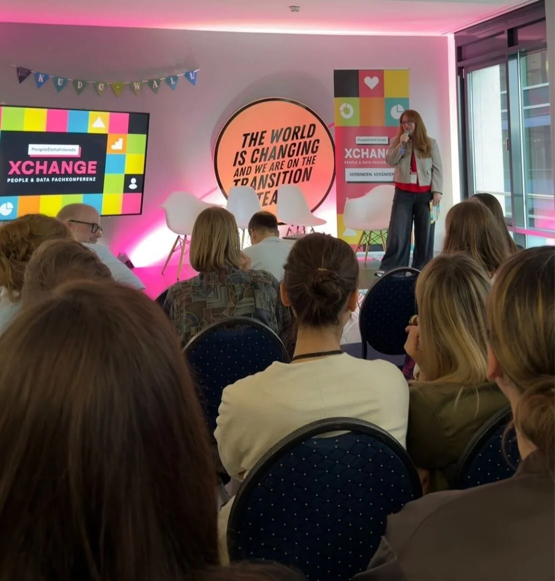 Eine Frau hält eine Rede auf einer Konferenz mit dem Thema "XCHANGE" vor einem Publikum. Im Hintergrund sind bunte Poster und ein Bildschirm mit dem Konferenznamen. Das Publikum hört aufmerksam zu.