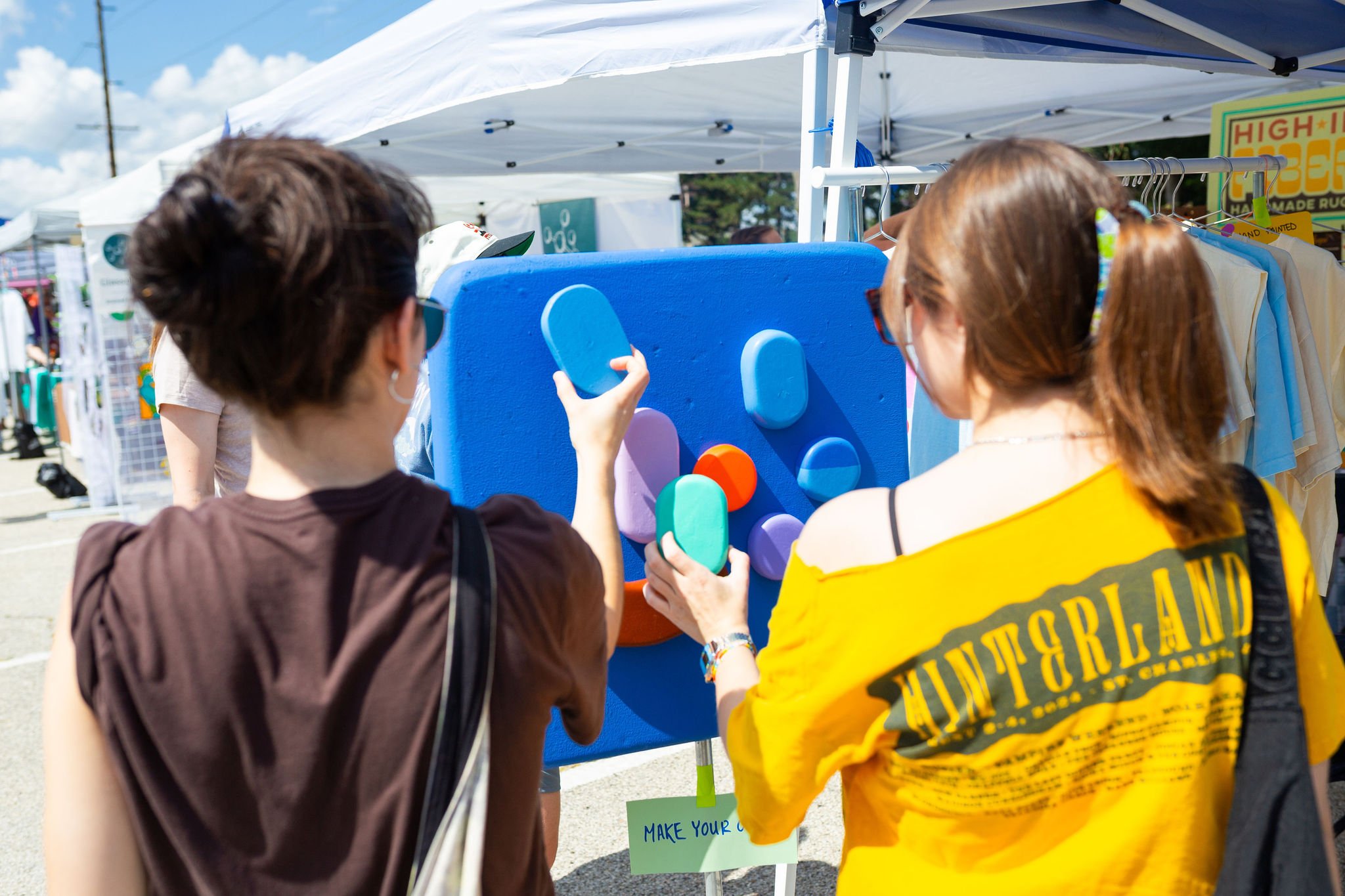 Three people at an outdoor event interacting with a colorful wall display featuring large foam shape pieces, with tents and vendor signs in the background.