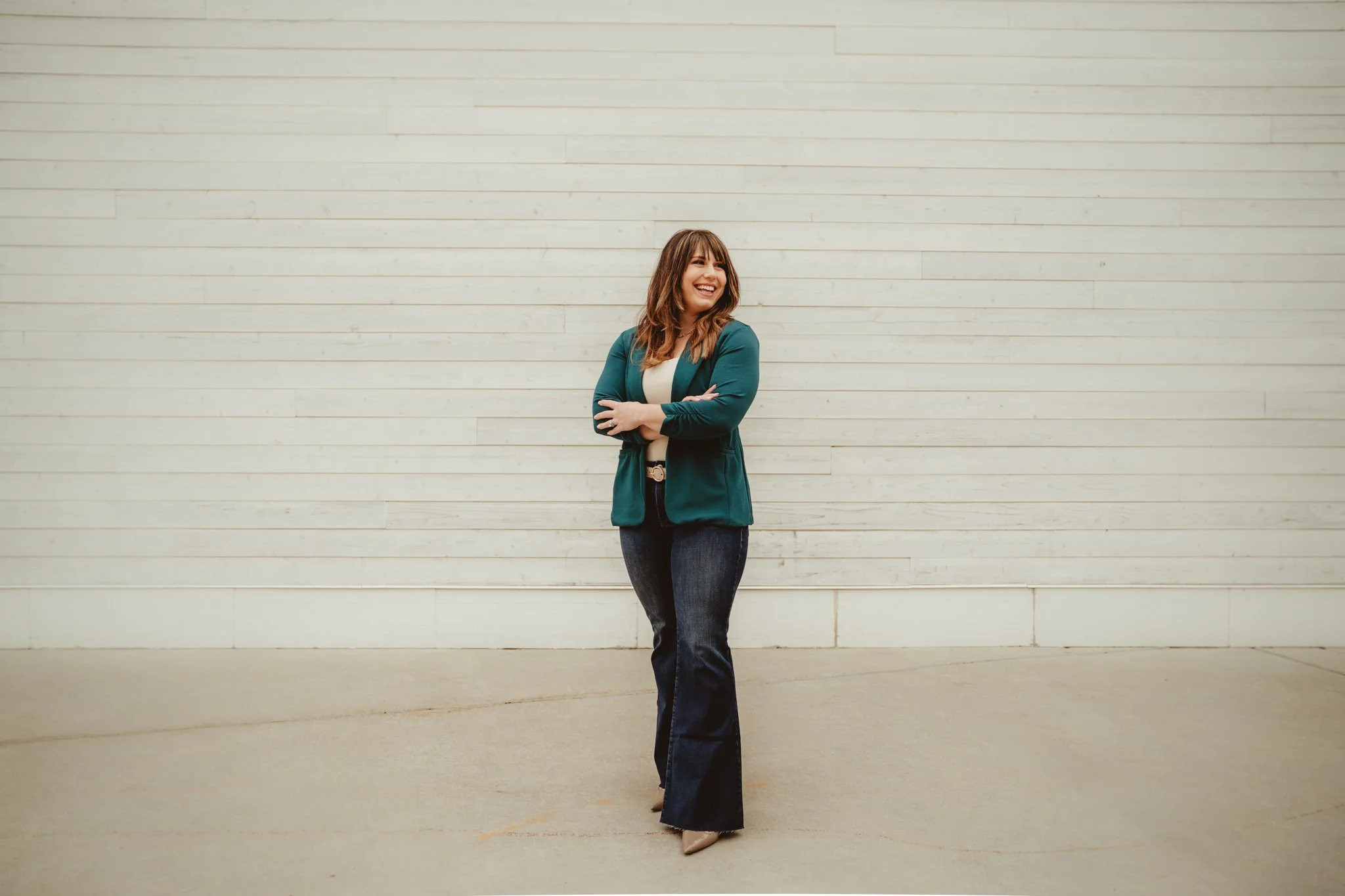 Woman standing in front of a beige wall, smiling with arms crossed, wearing a teal blazer, white top, dark jeans, and beige high heels.