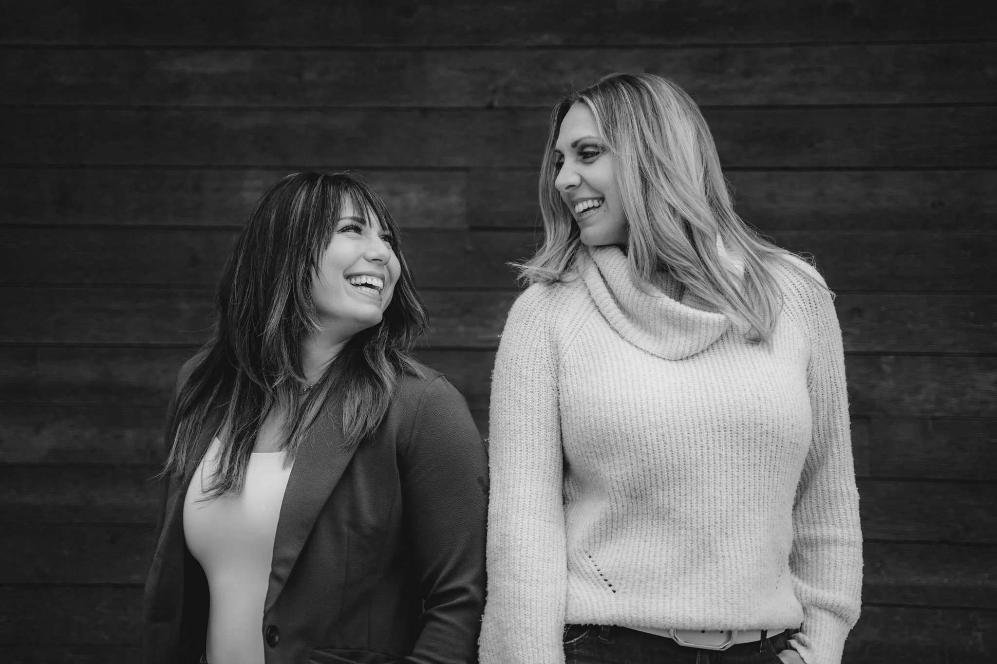 Two women smiling and looking at each other outdoors, standing in front of a dark wooden wall, in black and white.