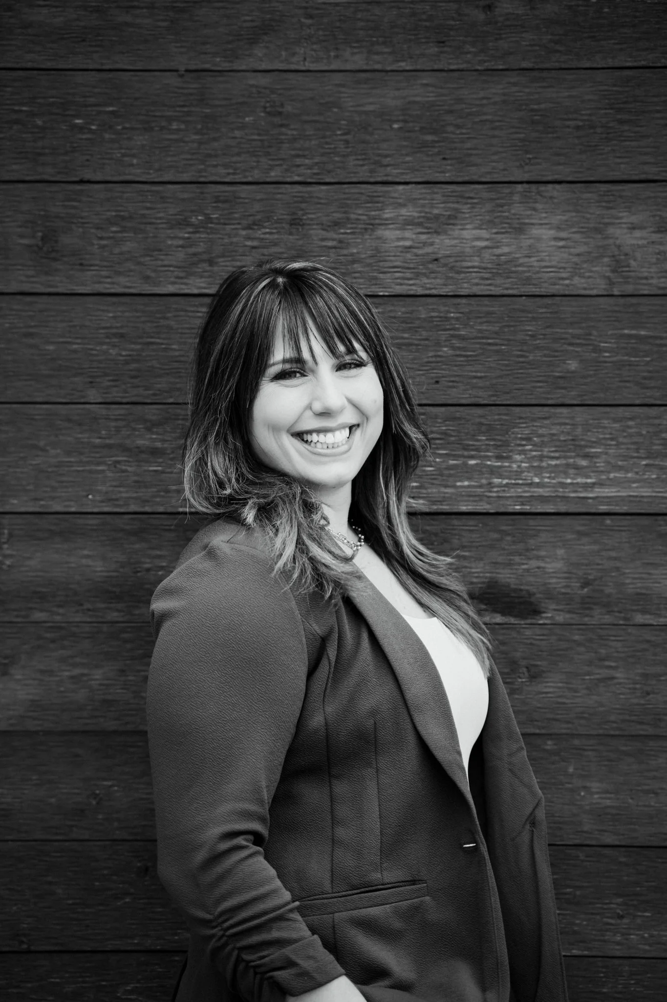 A smiling woman with shoulder-length hair wearing a blazer, standing against a wooden wall.