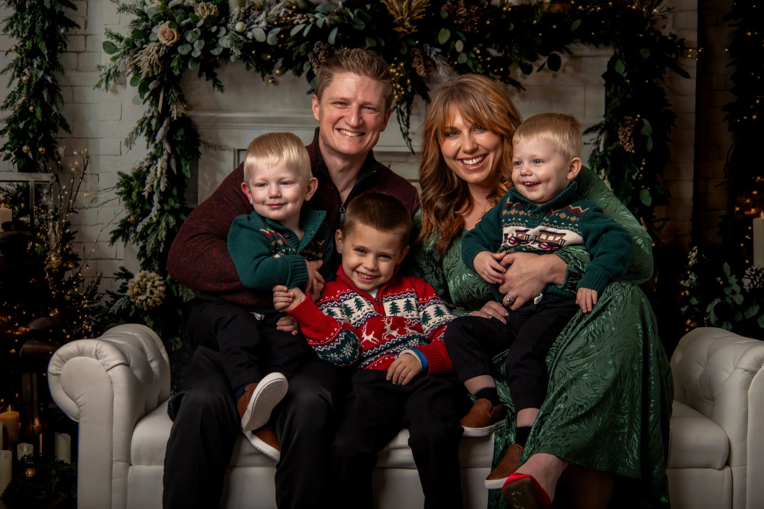 A family of six sitting on a white sofa in front of a decorated fireplace, celebrating Christmas. The group includes two adults and four children, all smiling and dressed in festive clothing. The background features Christmas tree decorations, candles, and a floral arrangement.