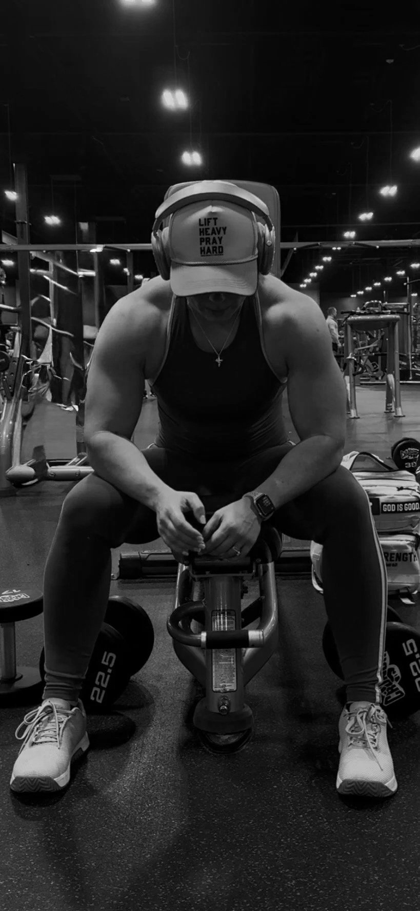 A person sitting on a workout bench in a gym, wearing a cap that reads 'LIFT HEAVY PRAY HARD', a tank top, headphones, and a wristwatch, surrounded by gym equipment.