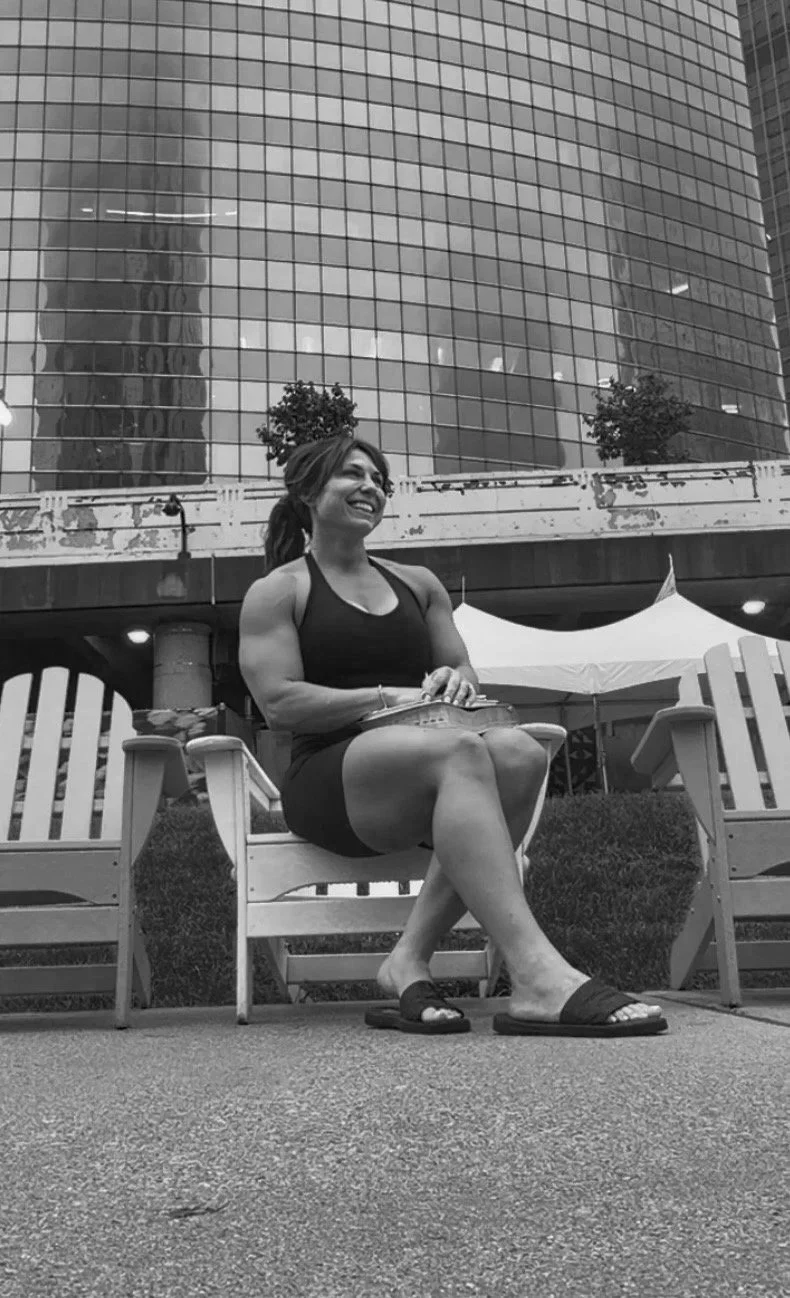 A woman in a sleeveless black dress and slide sandals sitting on a garden bench with her legs crossed, smiling, with a large office building with glass windows and a tent in the background.