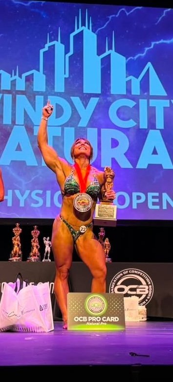 Female bodybuilder on stage at a competition, holding a trophy and wearing a medal around her neck. She is raising one arm with her index finger pointing up, smiling. Behind her is a large screen with a city skyline graphic and text that reads 'INDY CITY LAURA' and 'PHYSIQUE OPEN.' There are trophies and awards on the stage.