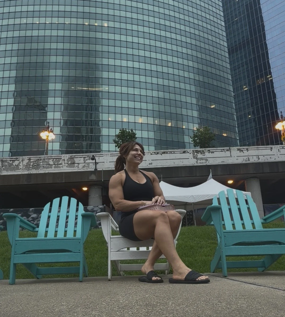 A woman in black athletic wear sitting on a white chair, smiling, with two blue chairs on either side, outdoors in front of a modern glass building.