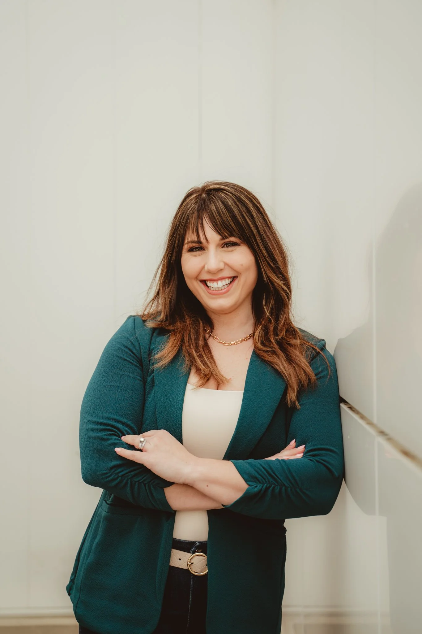 A woman with brown hair, smiling, wearing a teal blazer and black pants, standing with arms crossed, against a plain background.