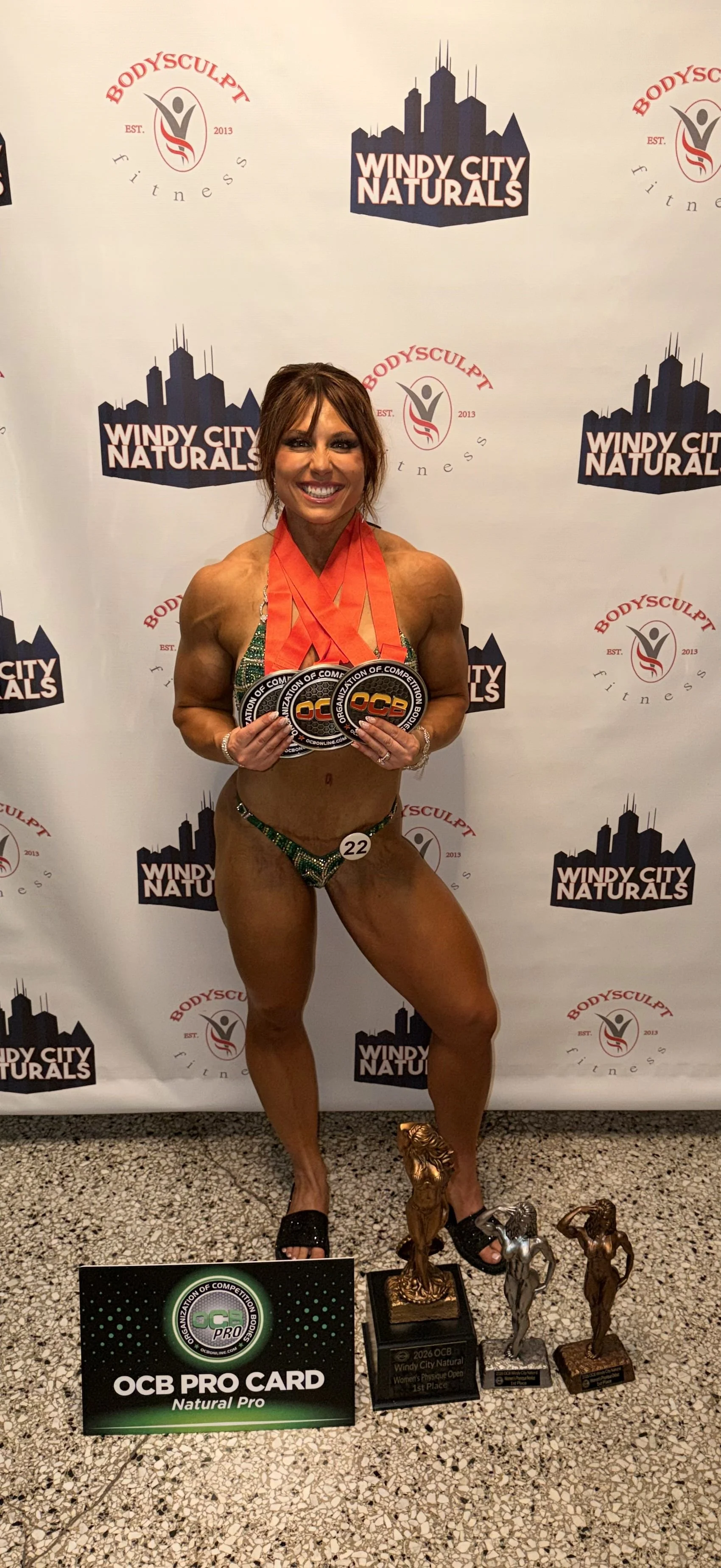 Female bodybuilder in green and black bikini holding medals, standing in front of a step and repeat banner with logos for Windy City Naturals and BodySculpt Fitness, displaying trophies on the floor.