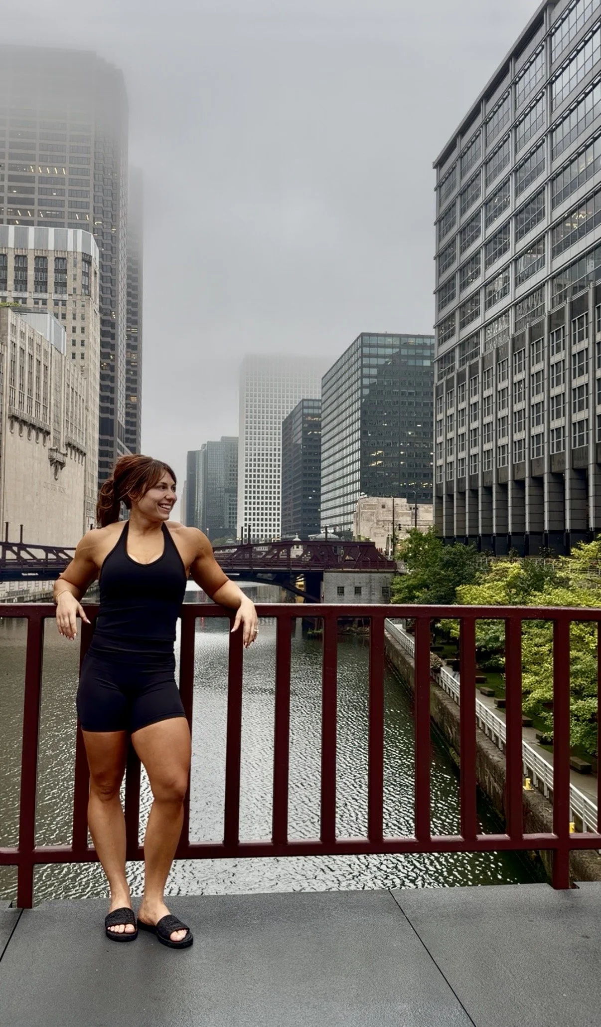 A woman in black athletic wear and sandals standing on a bridge in an urban area with tall office buildings and a river, under an overcast sky.