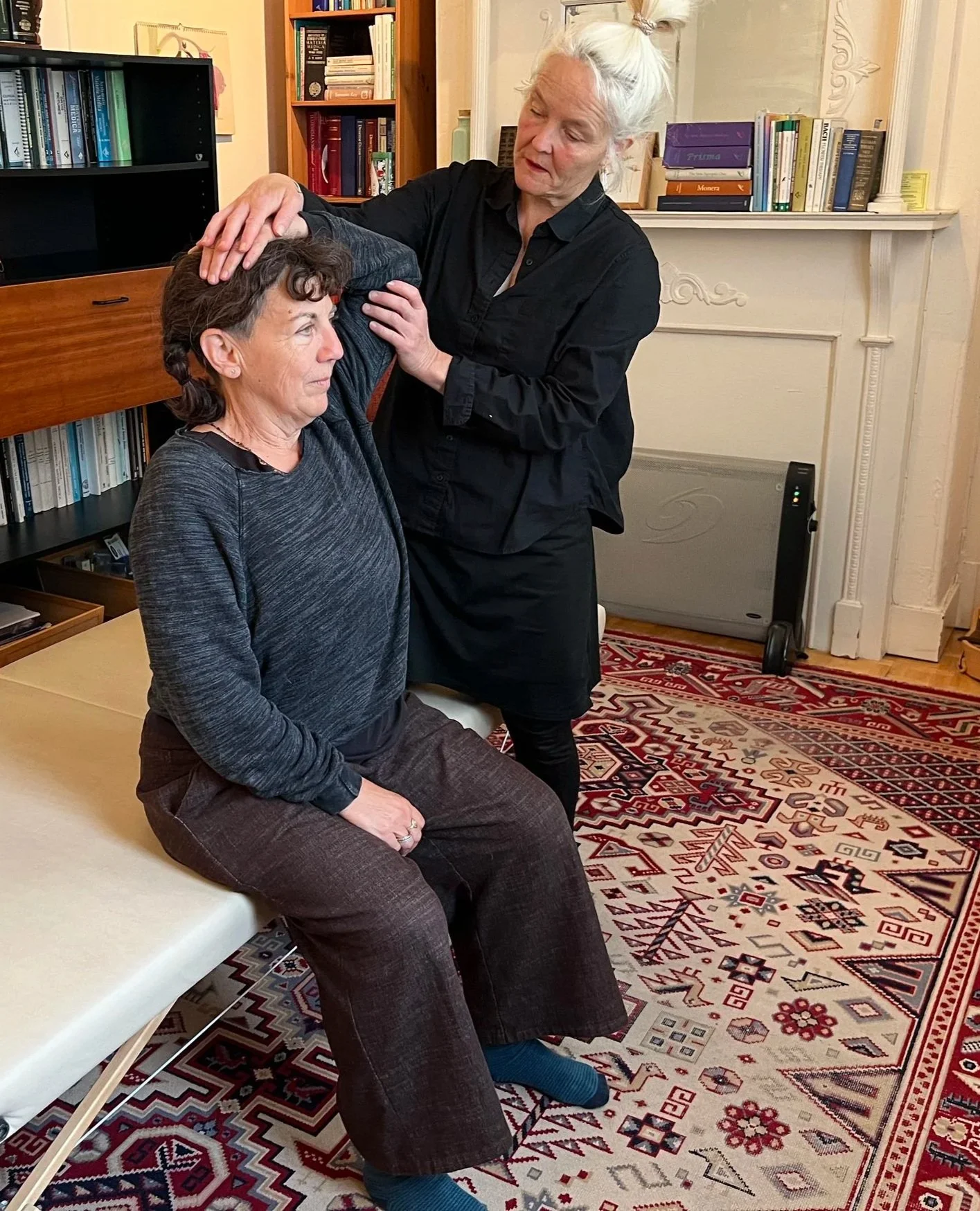 A middle aged woman with brown hair sitting on a bench with her eyes closed, receiving shoulder adjustment therapy from a woman standing beside her in a room with bookshelves and a patterned rug.