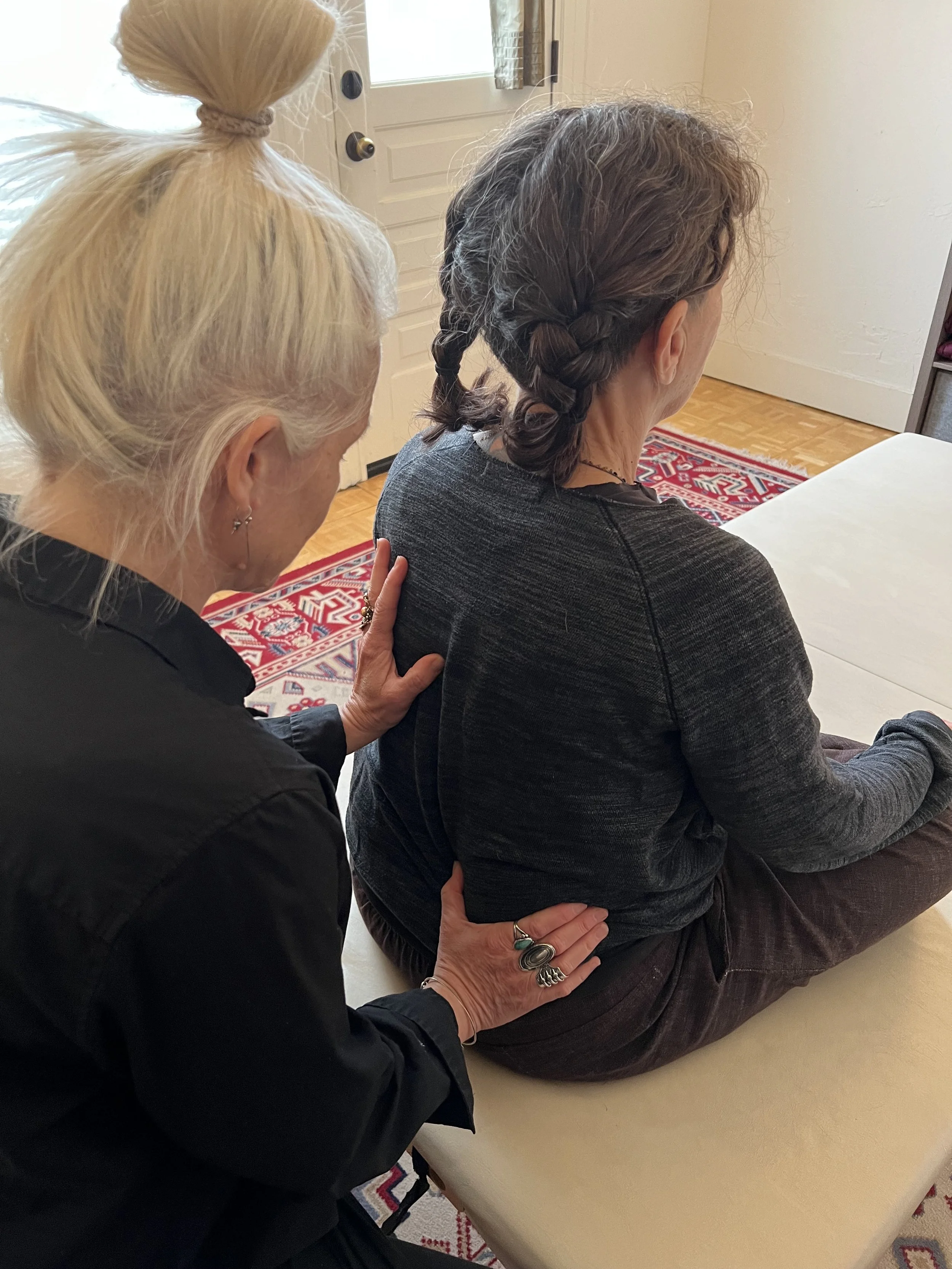 A woman with brown hair in braids sits on a massage table. A woman with platinum hair places her hands on the first woman's back