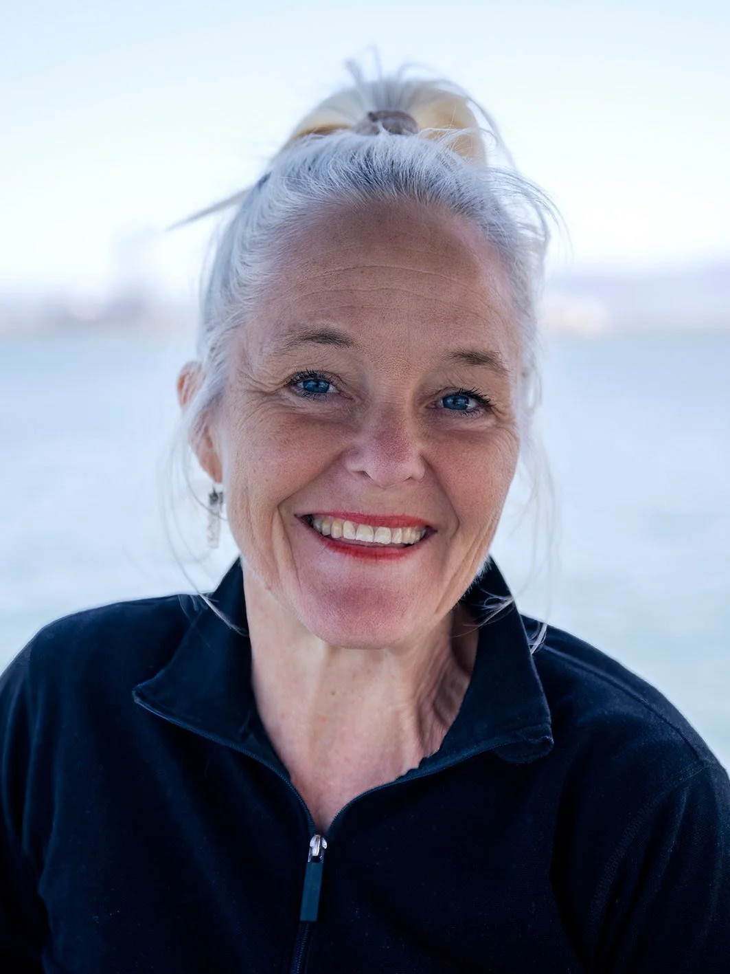 Close-up of smiling woman with blue eyes and platinum hair tied in a bun, wearing a black jacket, with a blurred body of water in the background.