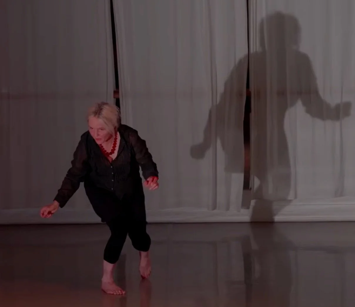 An woman dressed in black with a red necklace dancing barefoot in front of a curtain, with her shadow projected on the curtain.