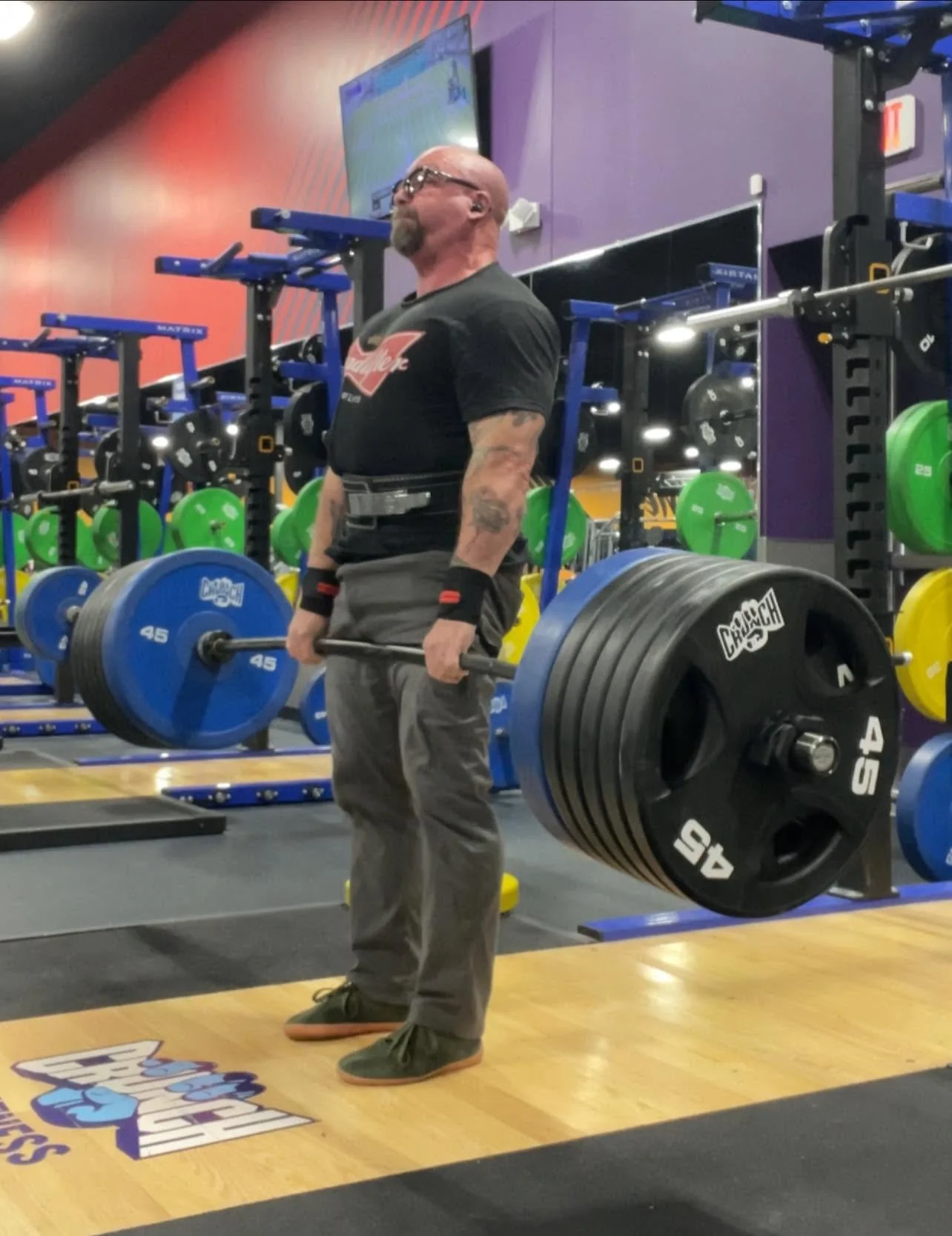 A man with tattoos and glasses lifting a heavy barbell with multiple weight plates in a gym.
